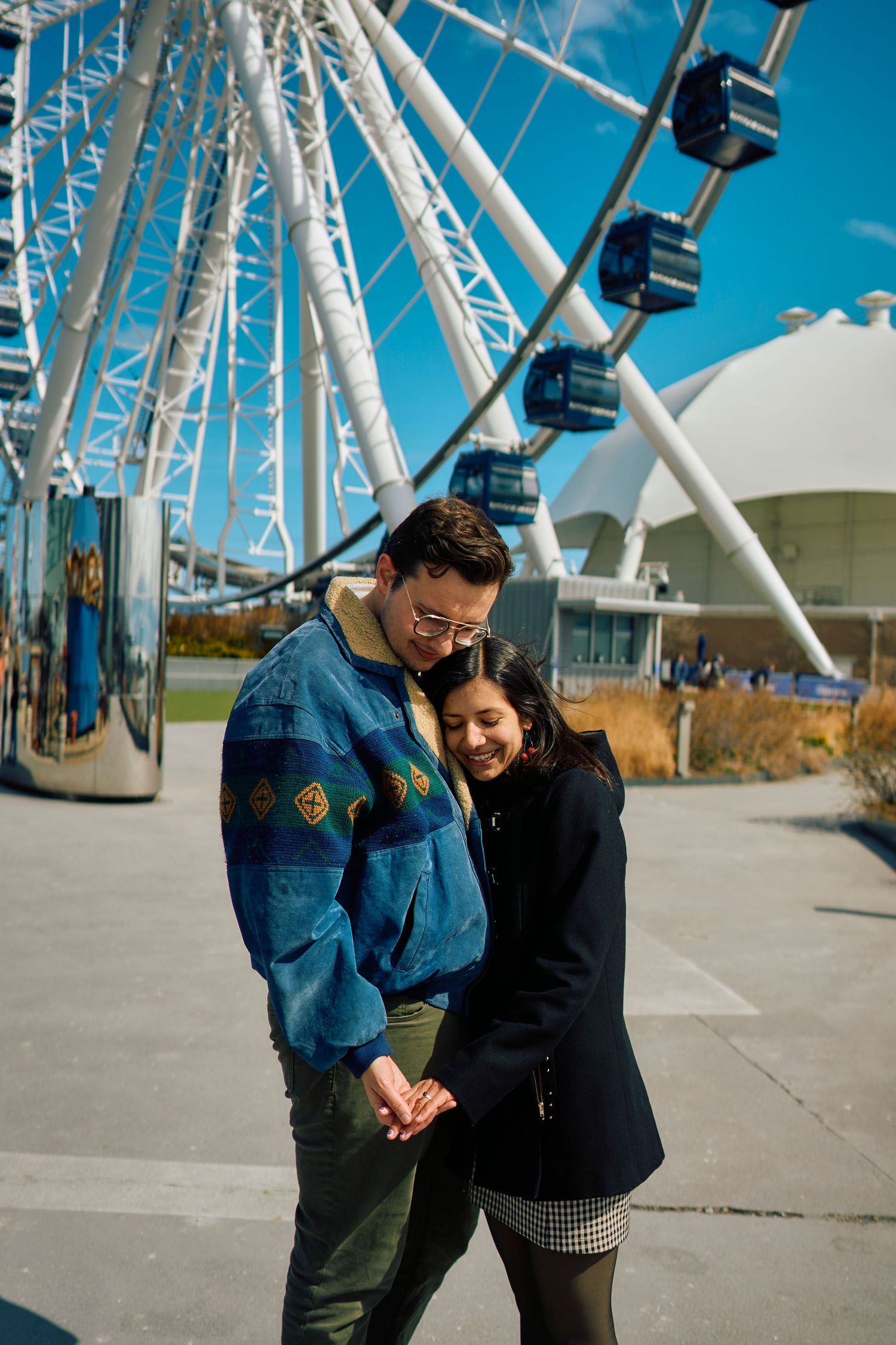 A couple embraces in front of a large Ferris wheel on a bright sunny day with blue skies.