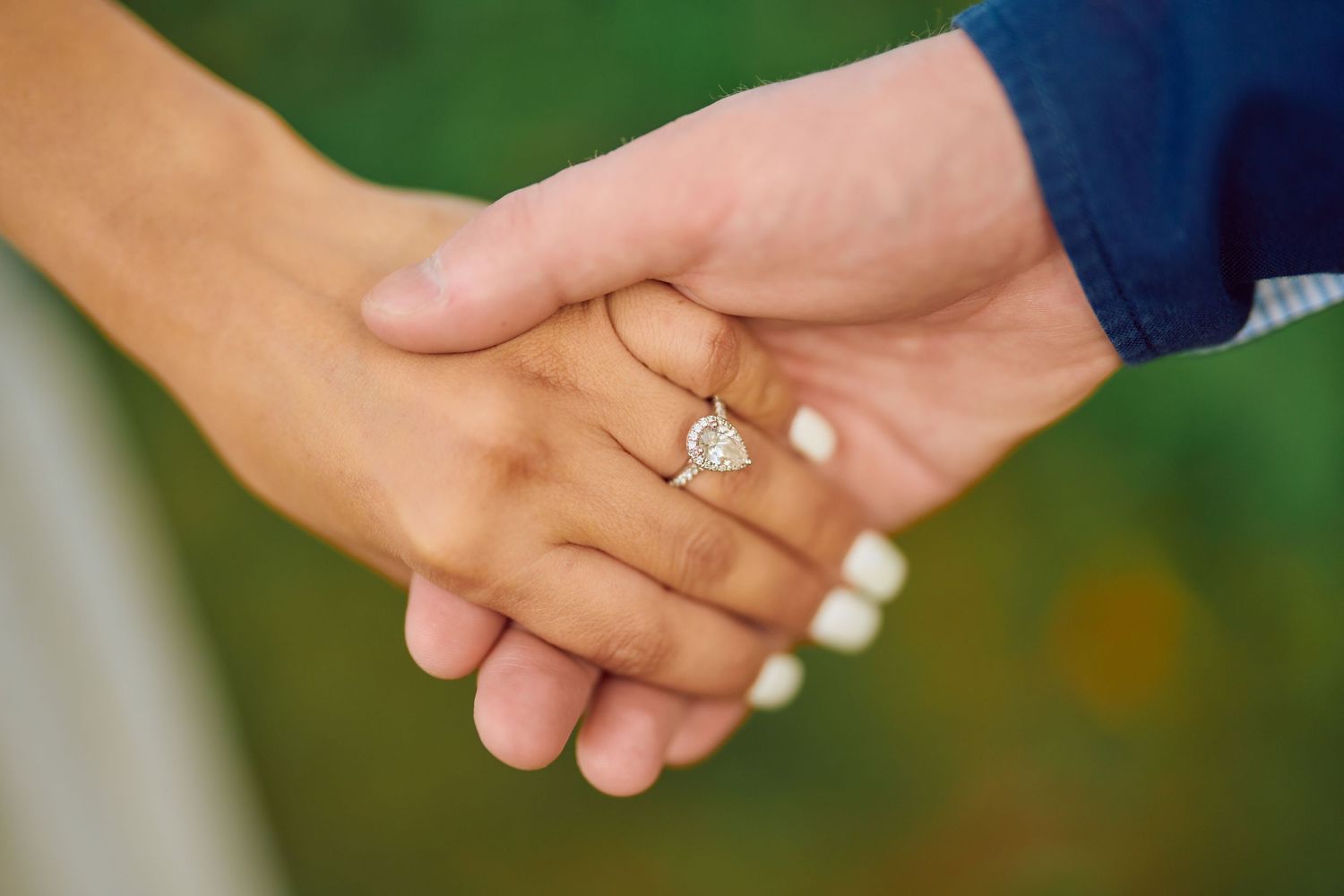 Close up of clasped hands showing a diamond engagement ring with a green blurred background.