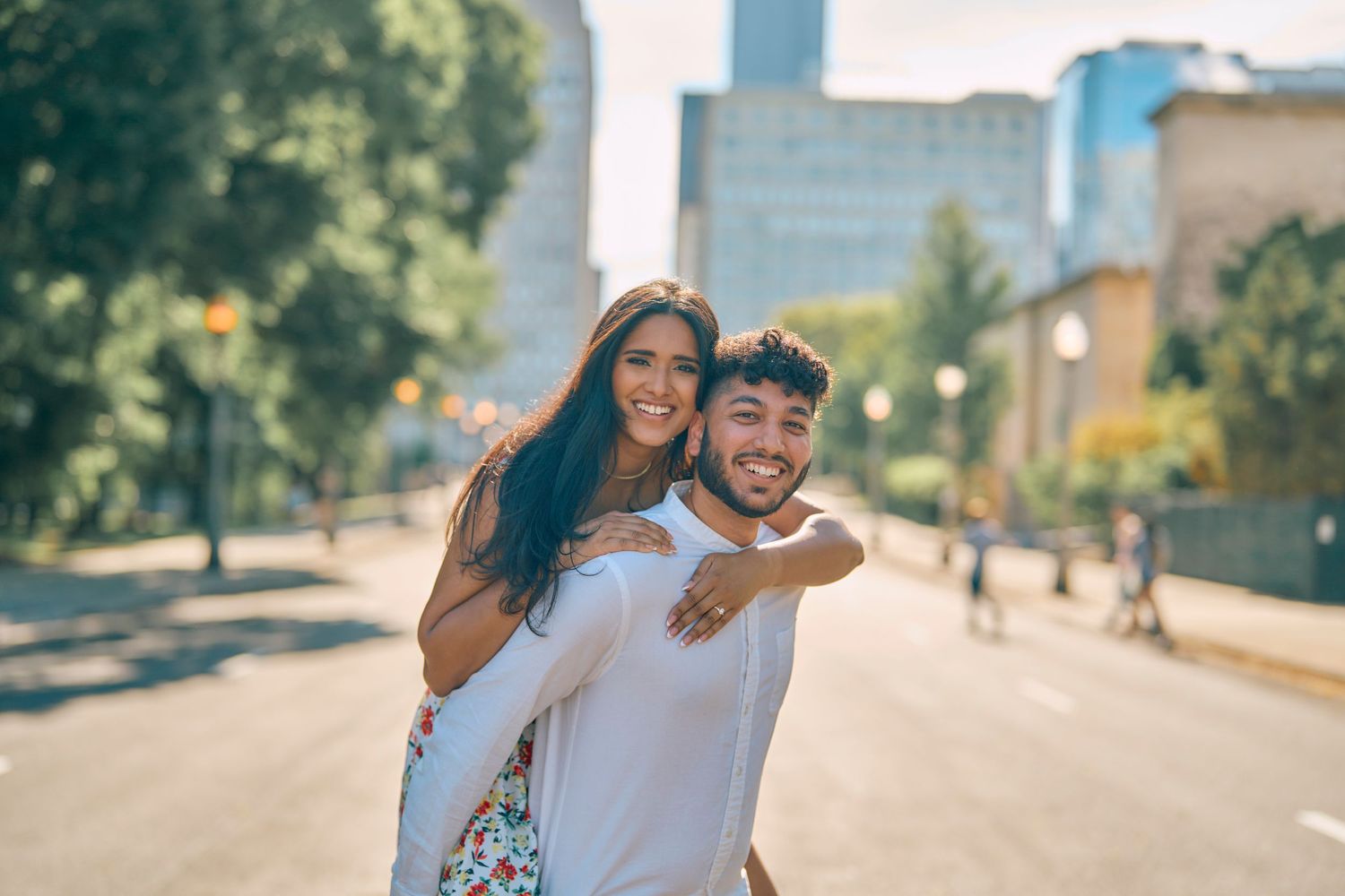 A happy couple enjoying a piggyback ride on a sunny urban street with trees and buildings in the background.