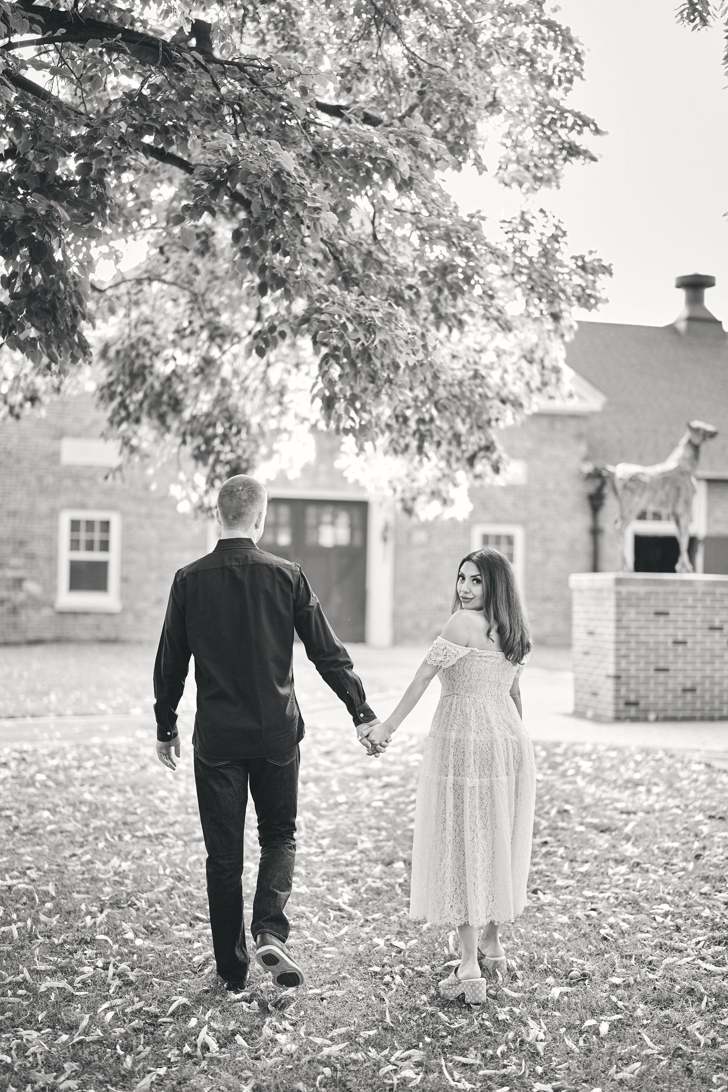 Romantic black and white photo of a couple walking hand in hand beneath a tree in front of a brick building.