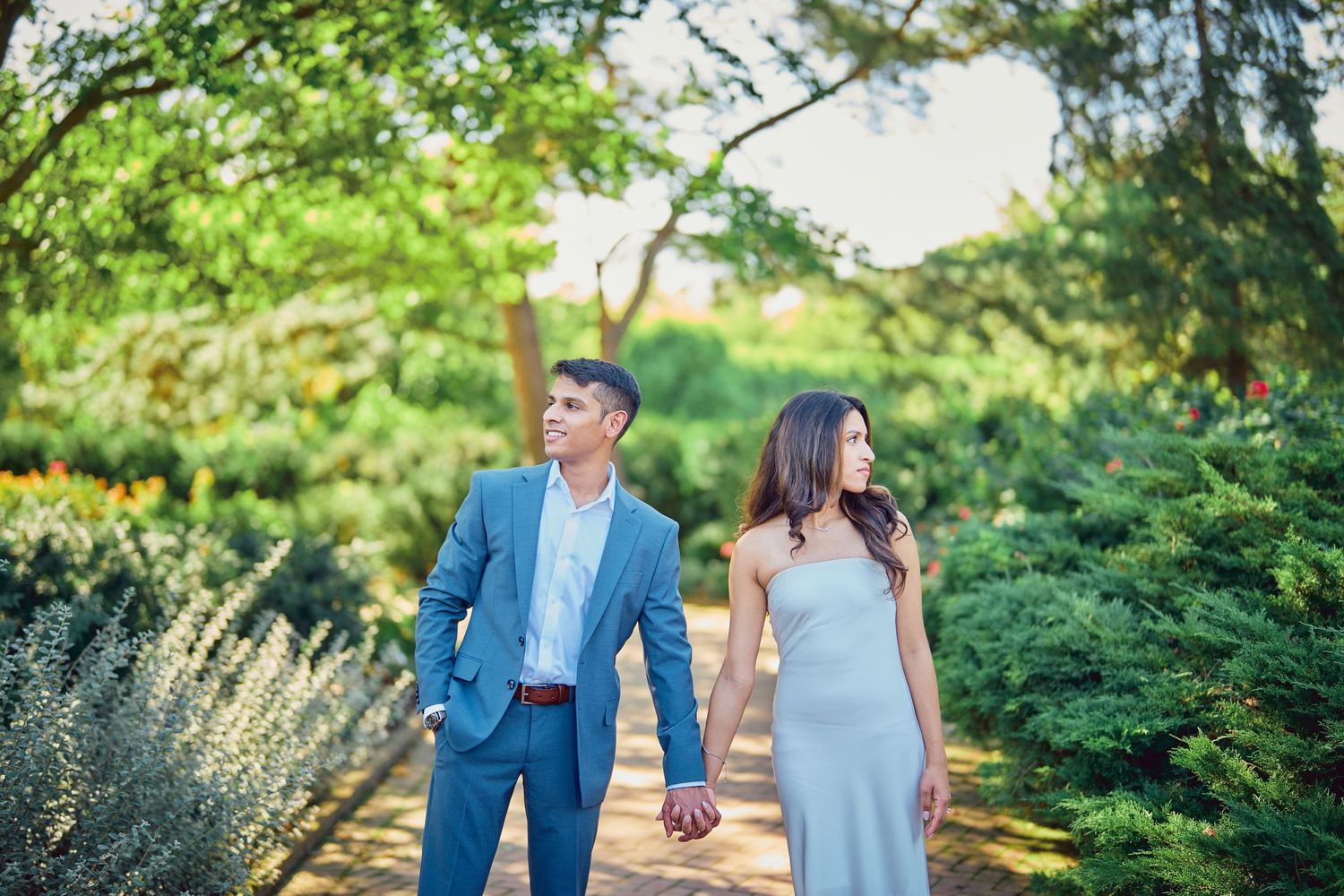 A couple in formal attire strolls hand-in-hand through a lush garden setting.