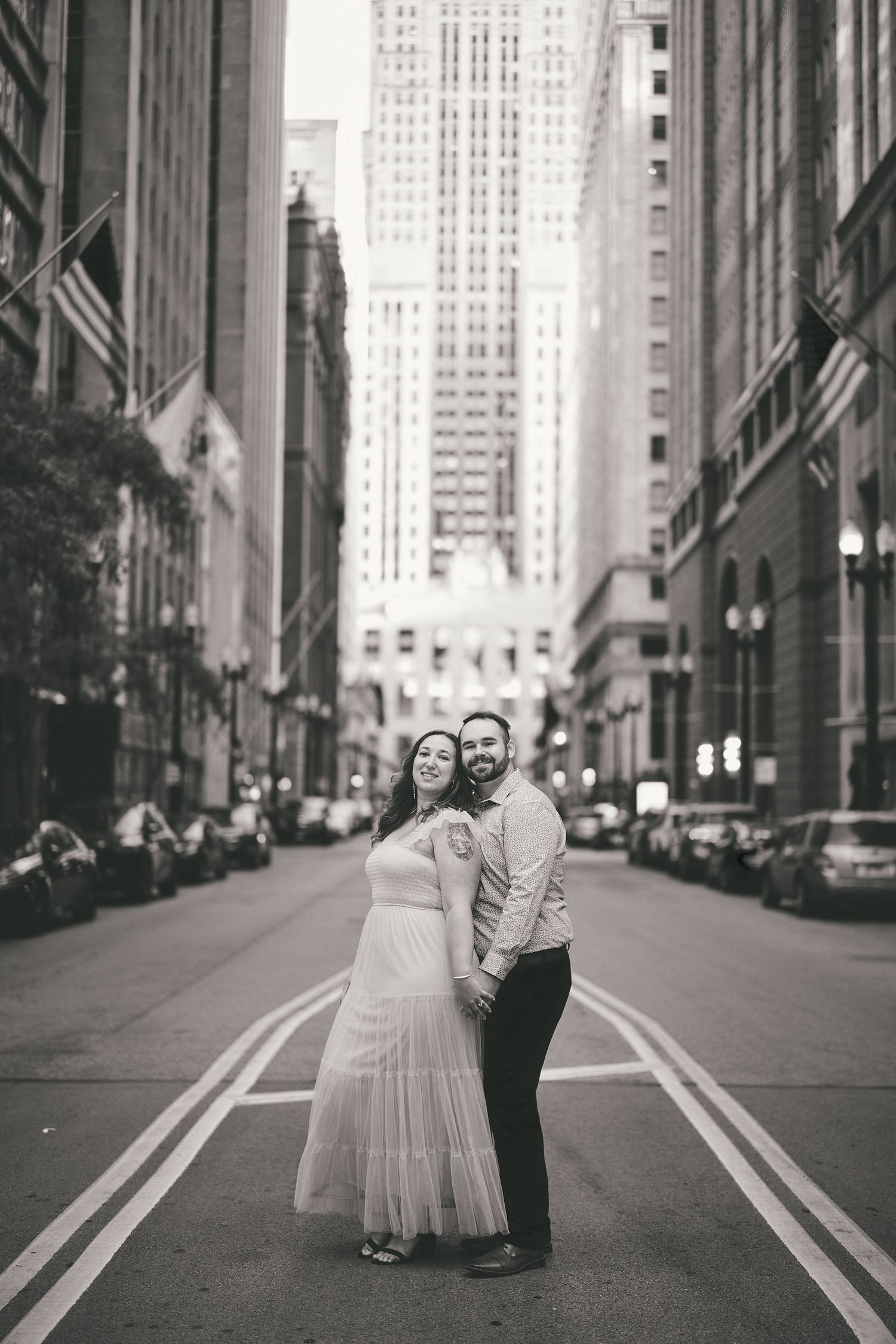 A black and white urban photo showing a couple embracing in the middle of a city street surrounded by tall buildings.
