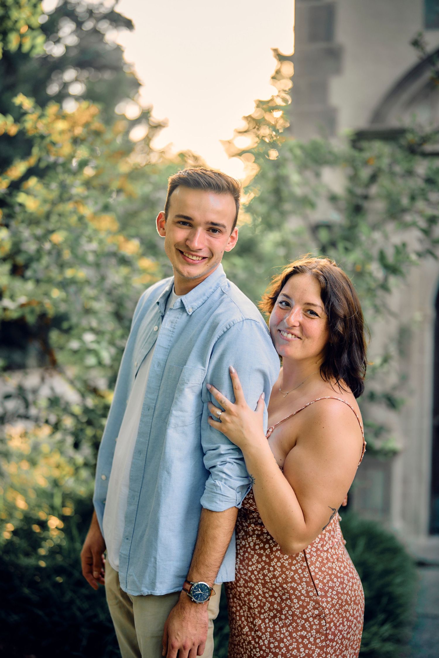 A couple poses together outdoors in casual summer attire with greenery and soft sunset lighting in the background.