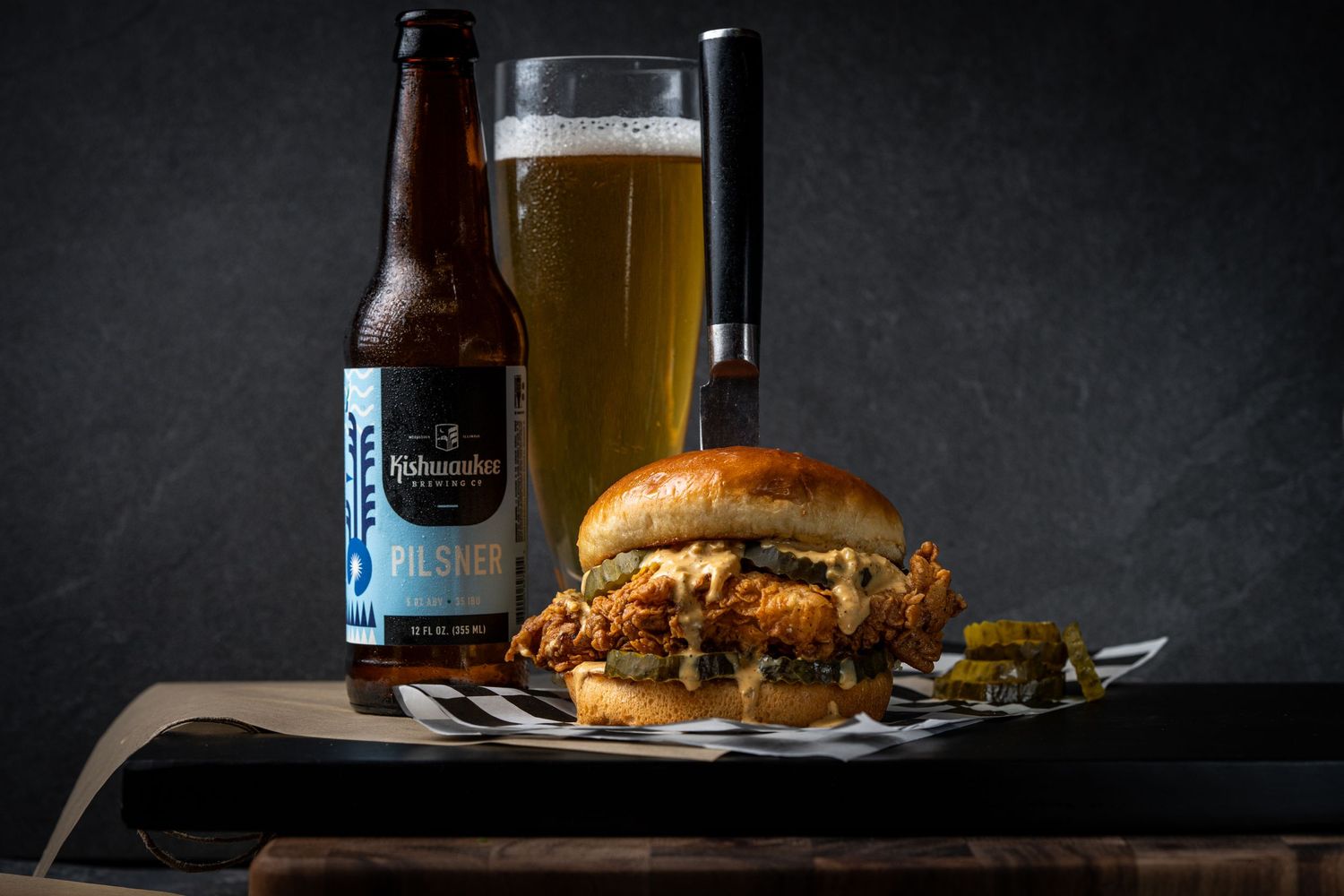 Craft beer bottle and glass beside loaded burger on black surface.