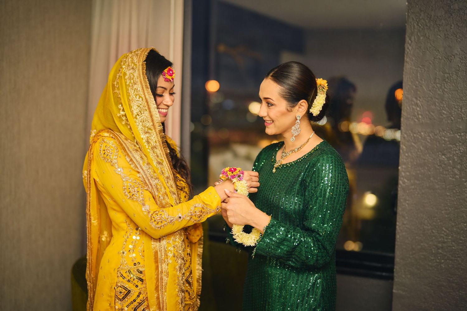 Two women in traditional South Asian attire, one in yellow and one in emerald green, share a joyful moment at a formal event.