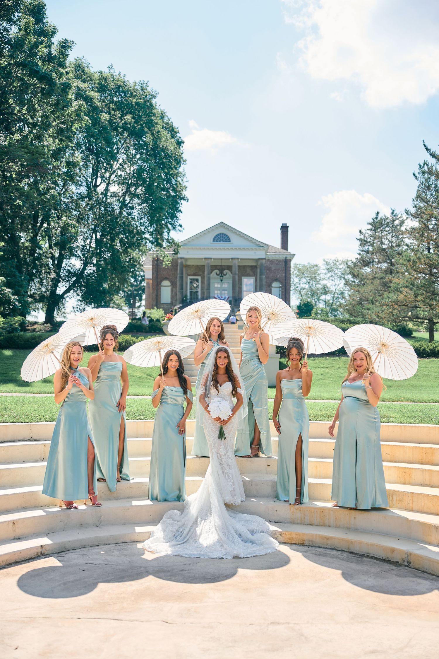 Wedding party with bridesmaids in mint green dresses holding white parasols pose on stone steps at elegant venue.