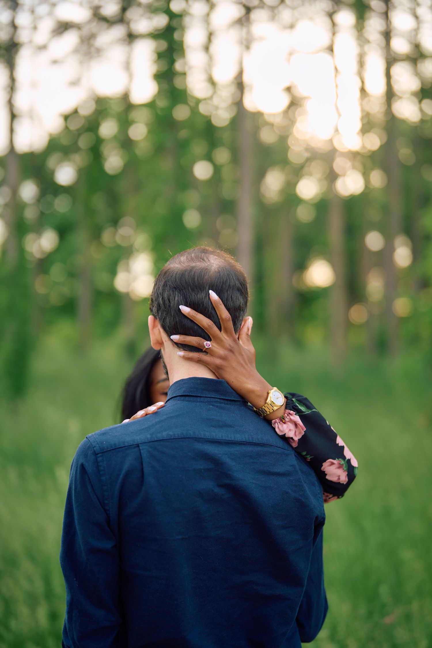 A couple embraces in a romantic pose against a forest backdrop with soft bokeh lighting at sunset.