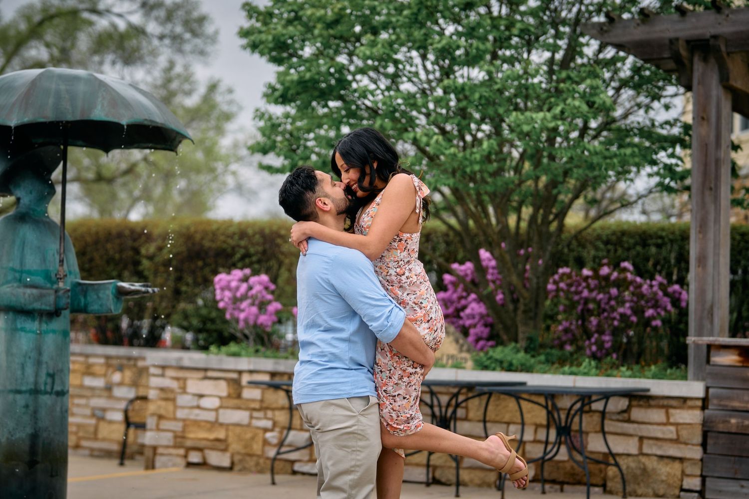 A romantic moment captured as a couple embraces in front of blooming azaleas while someone holds an umbrella.