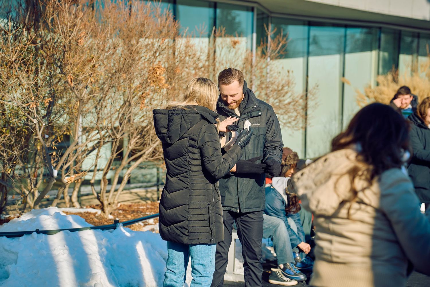 A couple wearing winter coats share a moment together outside a building on a sunny day.