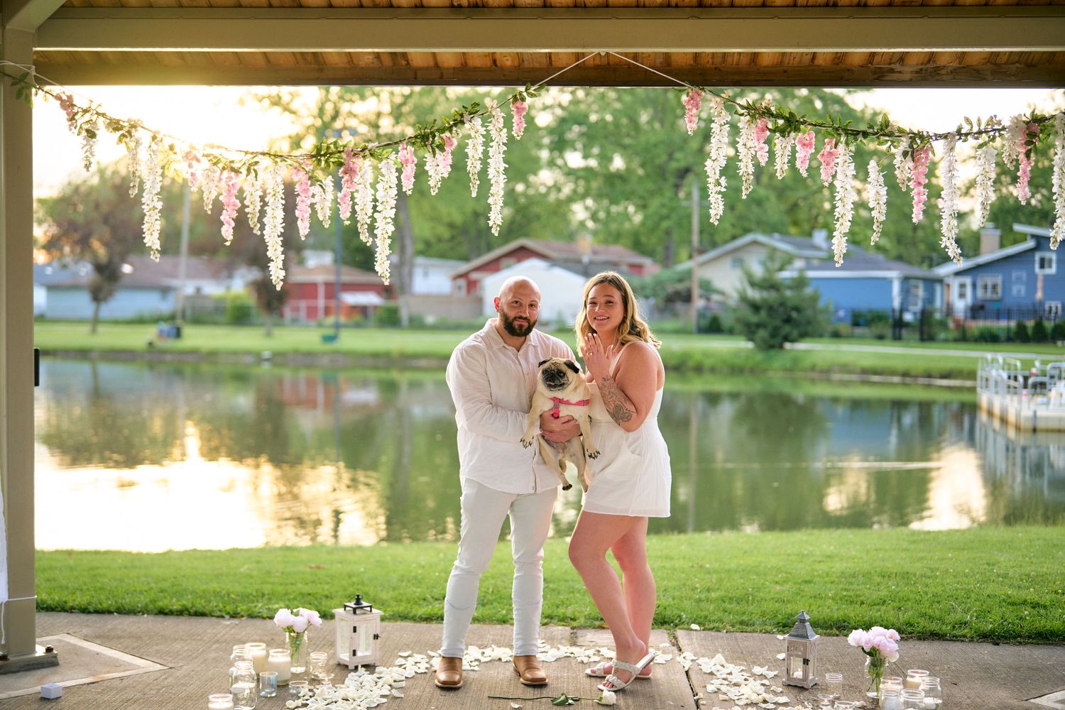 A couple stands under romantic floral gazebo by a serene lake during sunset with string lights and decorations.