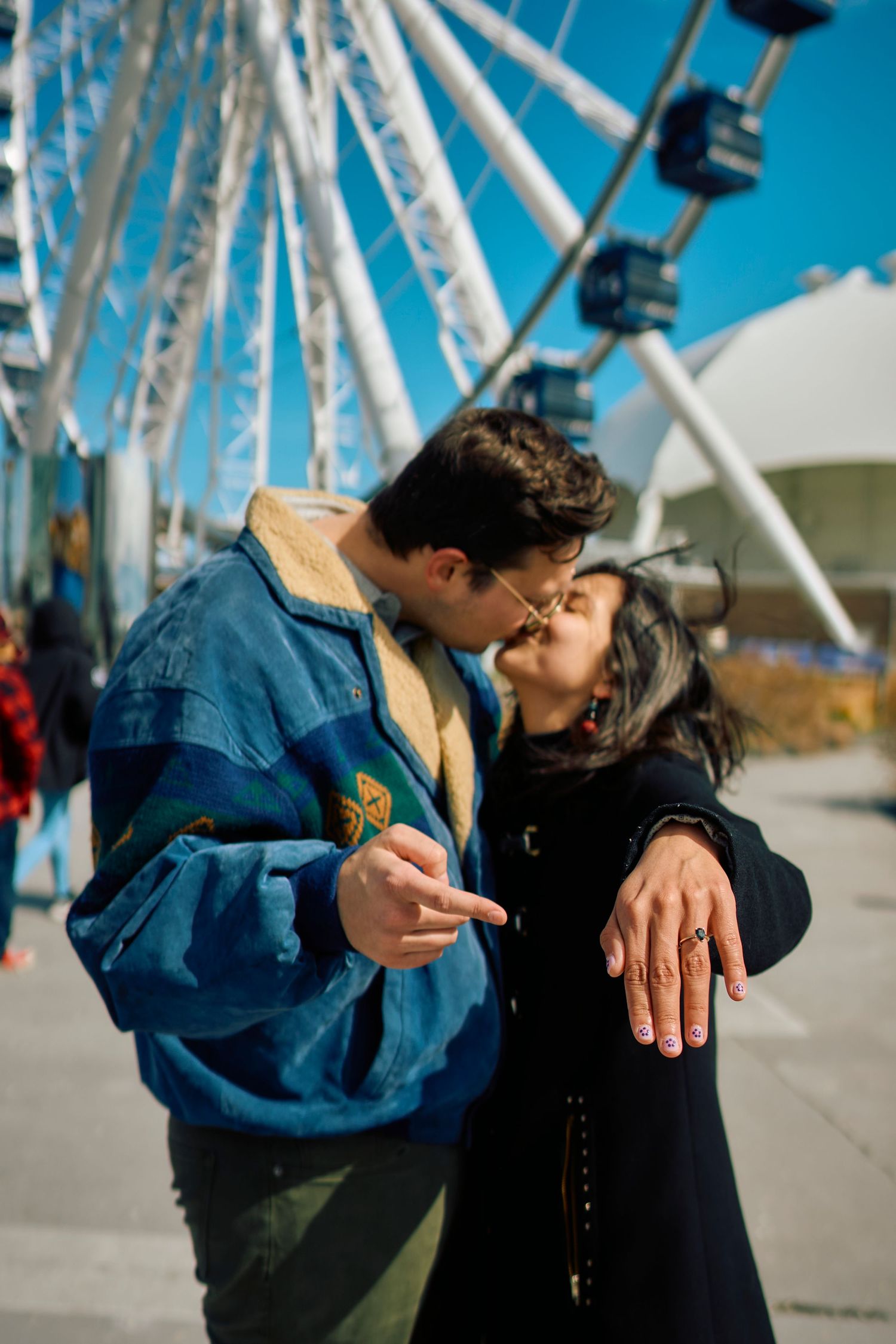 A romantic couple sharing a kiss in front of a ferris wheel on a sunny day with blue skies.