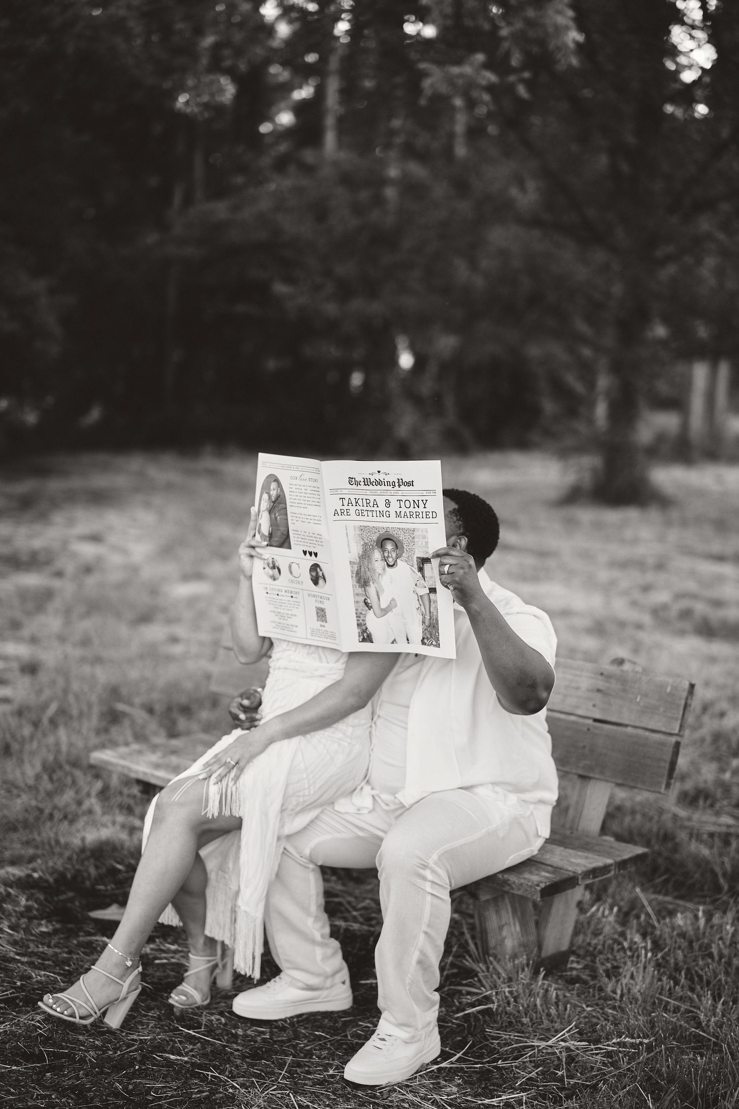 A couple in white outfits sits on a bench reading a newspaper together in a black and white photo.