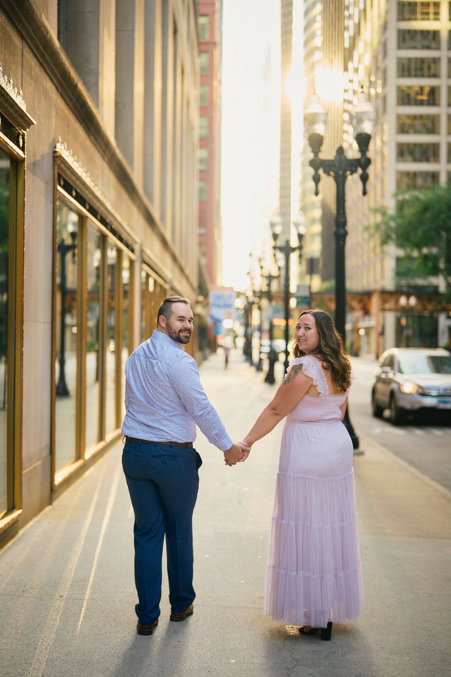 A couple holding hands while walking on an urban sidewalk with vintage lampposts and tall buildings in Chicago.
