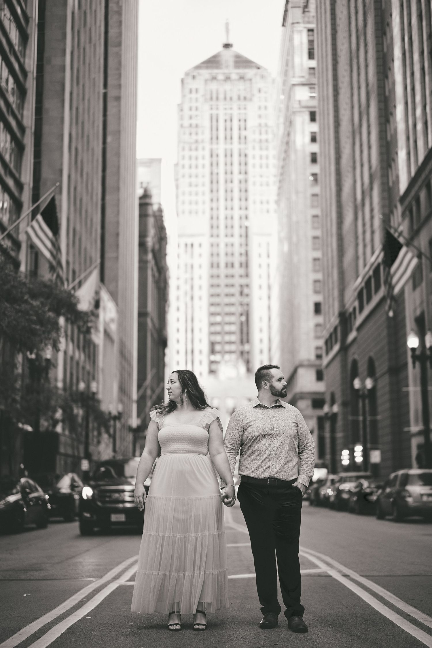 A couple stands together in a black and white photo on a Chicago street with tall buildings towering above them.