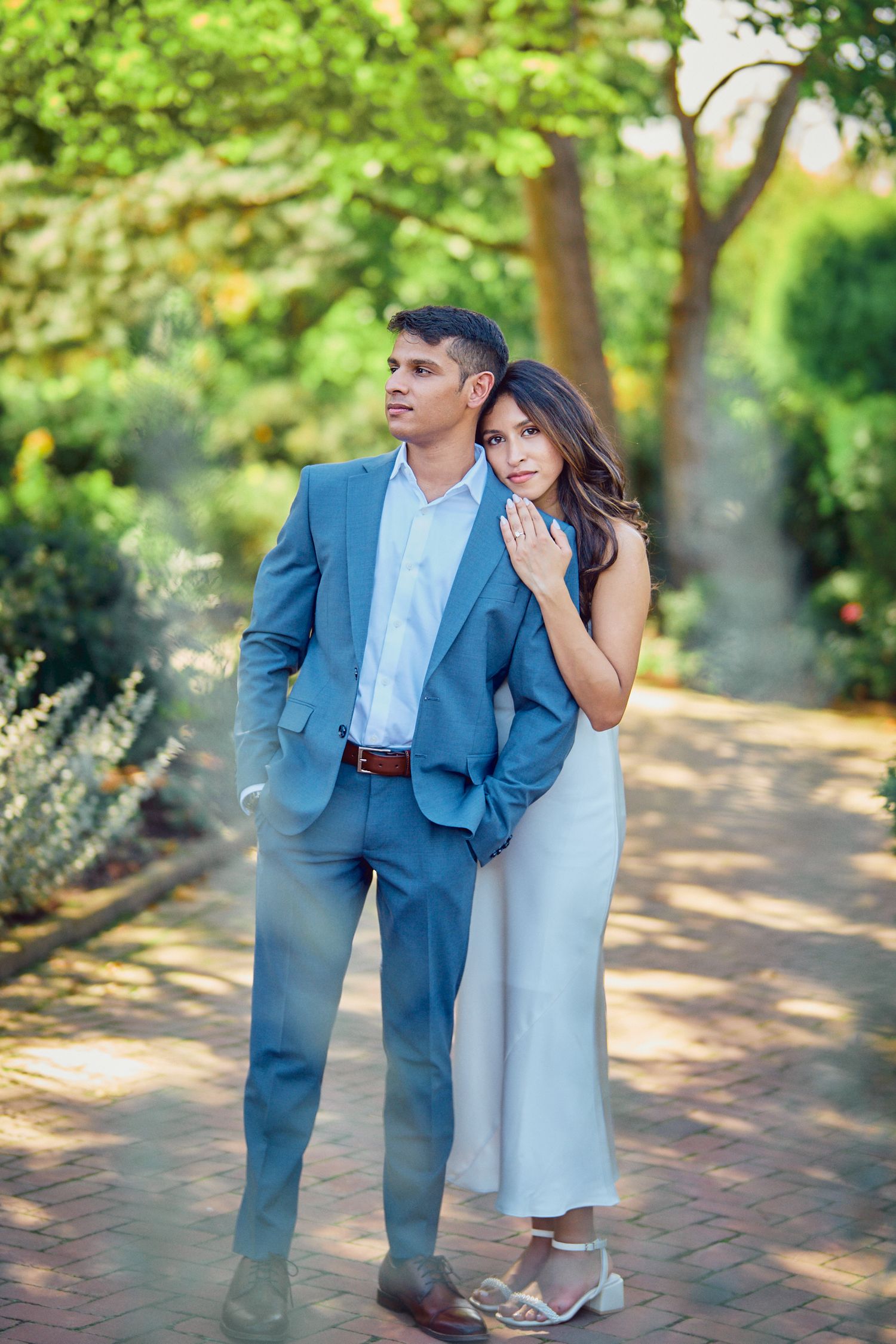 A romantic couple poses on a garden path wearing coordinated formal attire with blue and white tones.