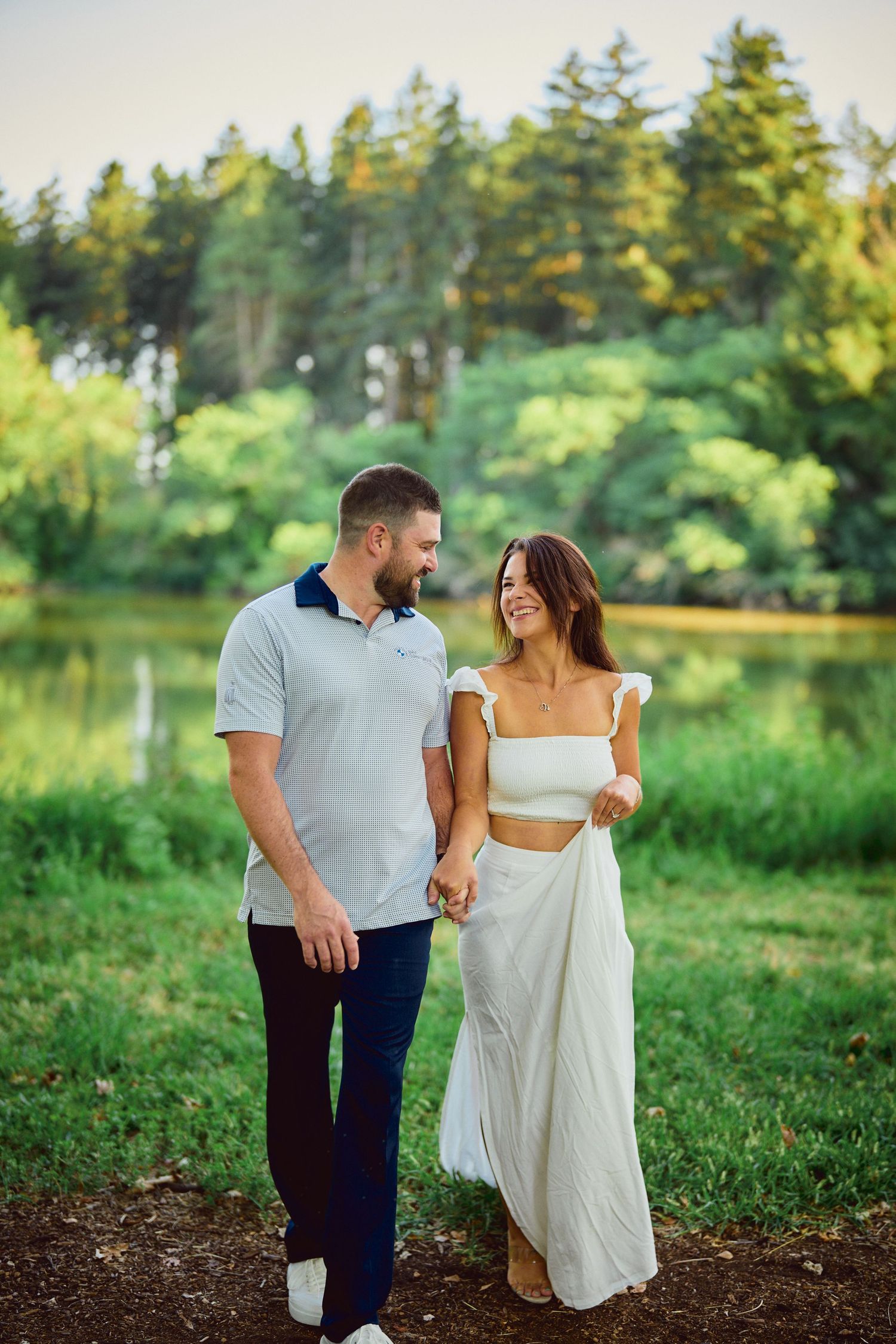 A couple takes a romantic stroll along a lakeside path surrounded by lush evergreen forest on a sunny summer day.