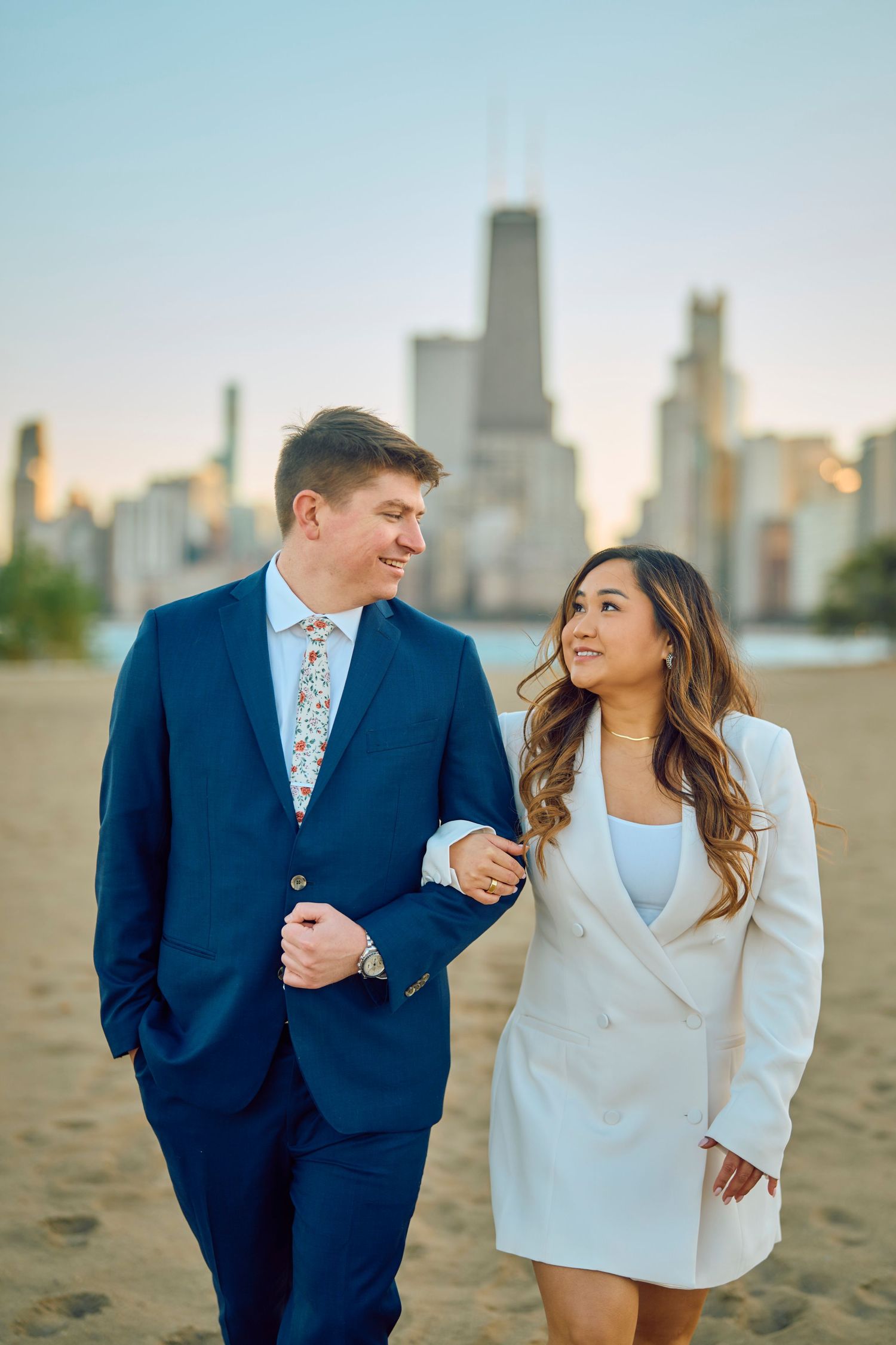 A well-dressed couple walking on a beach with Chicago skyline in the background at sunset.