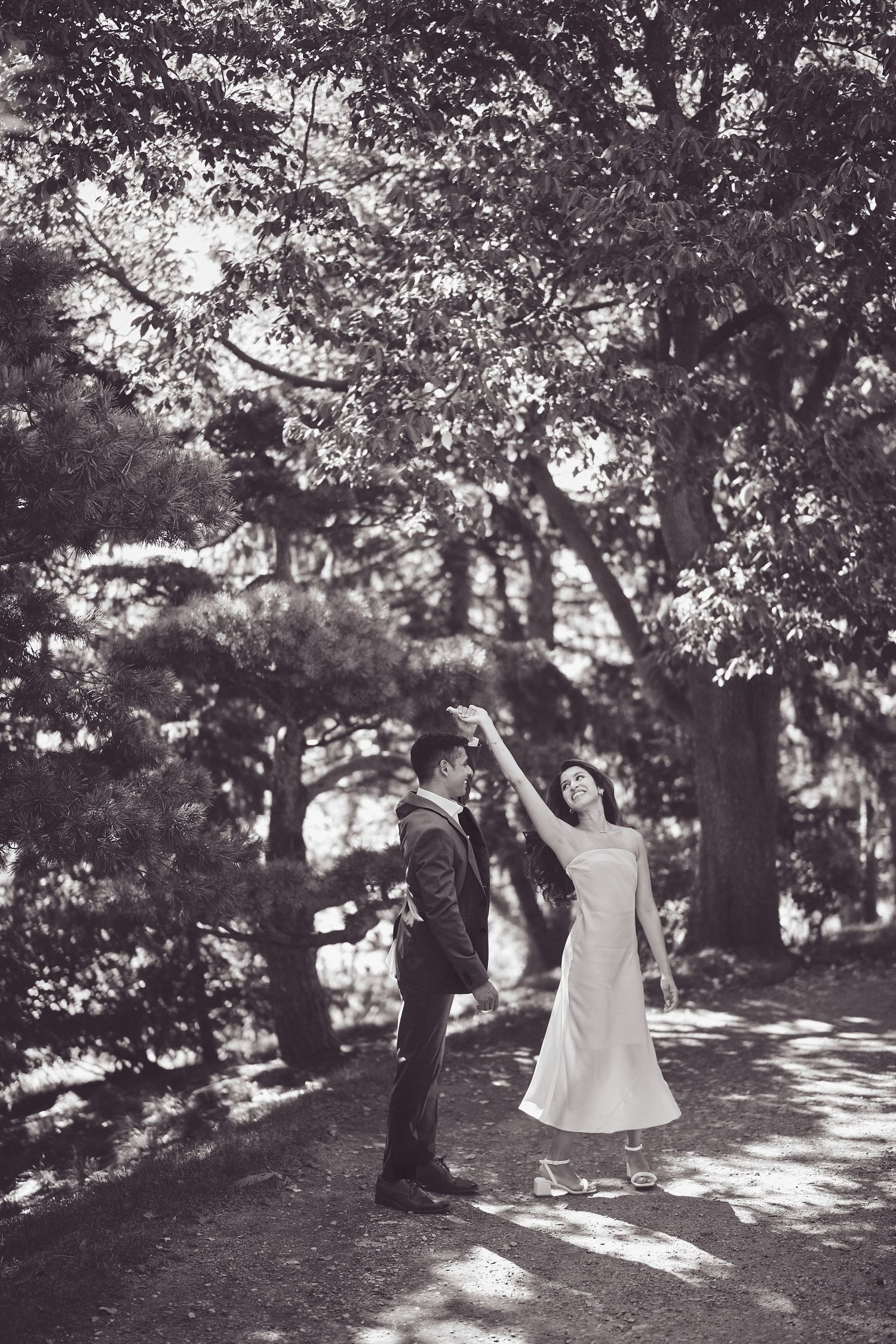 A couple dances together on a tree-lined path with dappled sunlight filtering through the branches.