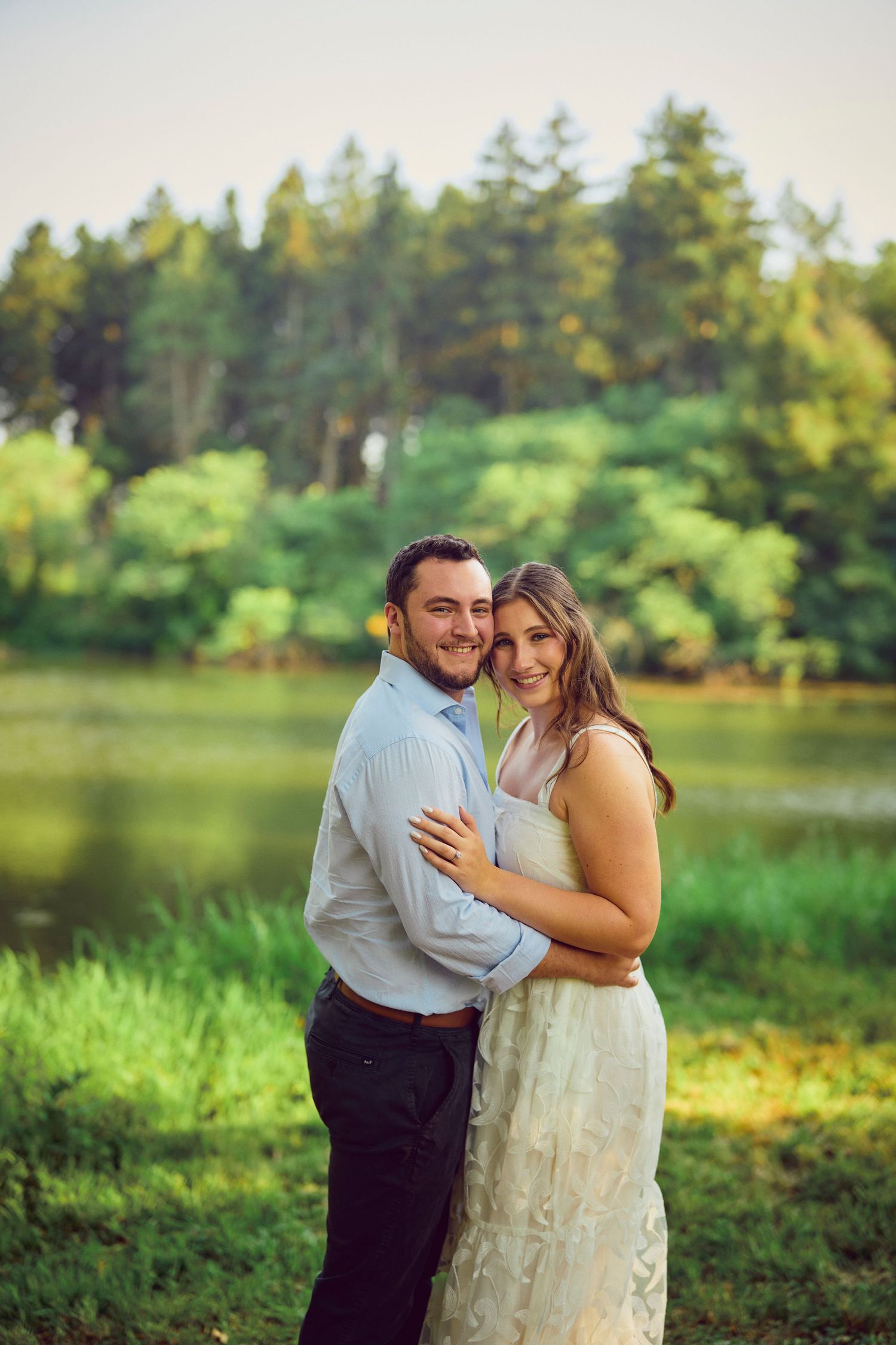 A couple embraces in front of a serene lake surrounded by vibrant green trees and foliage.