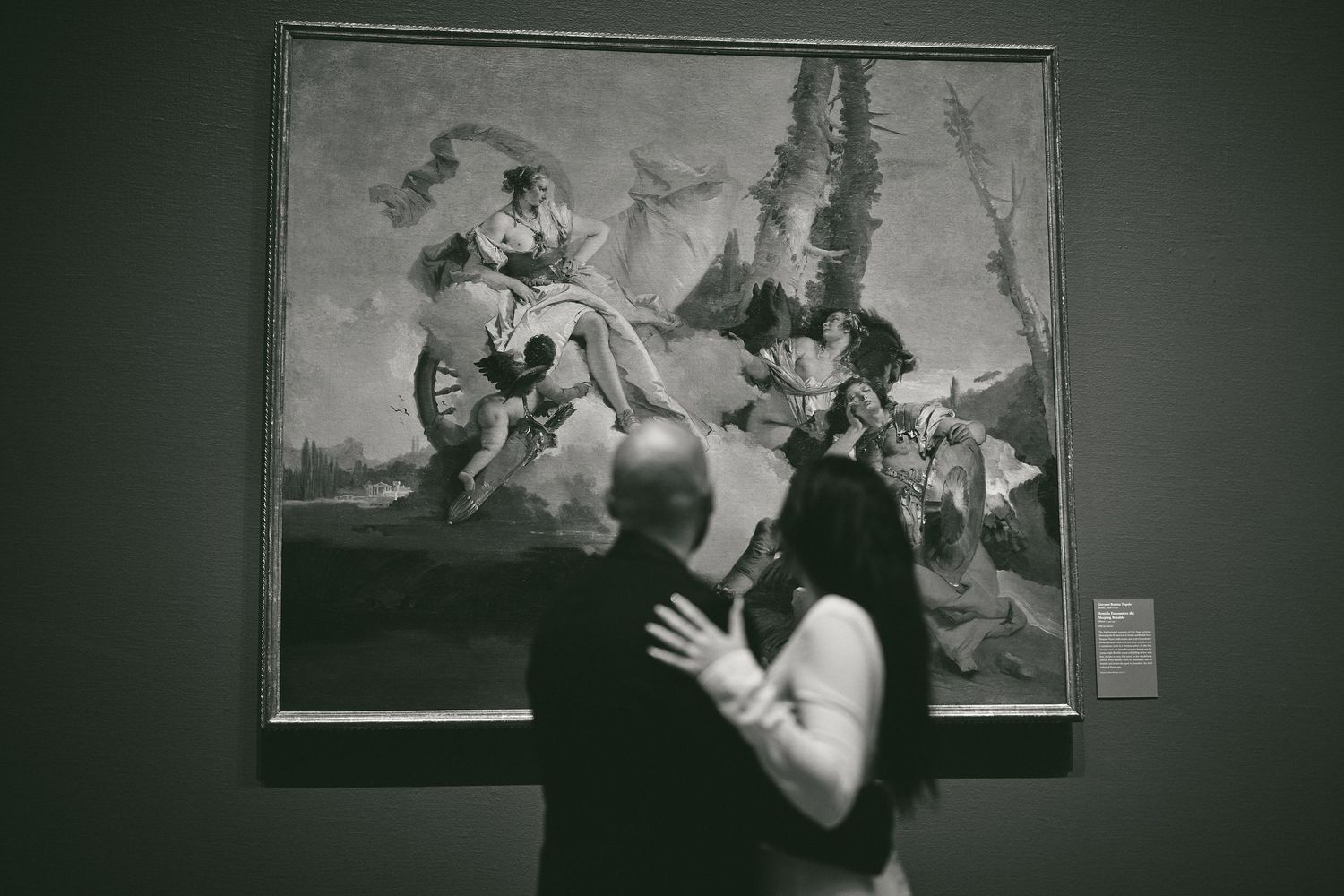 A person with their hands raised views a dramatic classical painting mounted on a gallery wall.