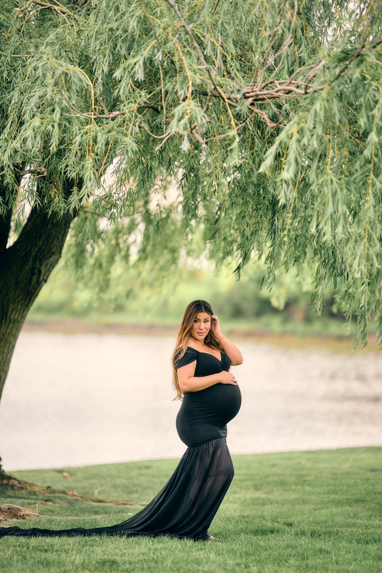 A graceful figure in a black flowing gown poses under a weeping willow tree beside a serene lake.