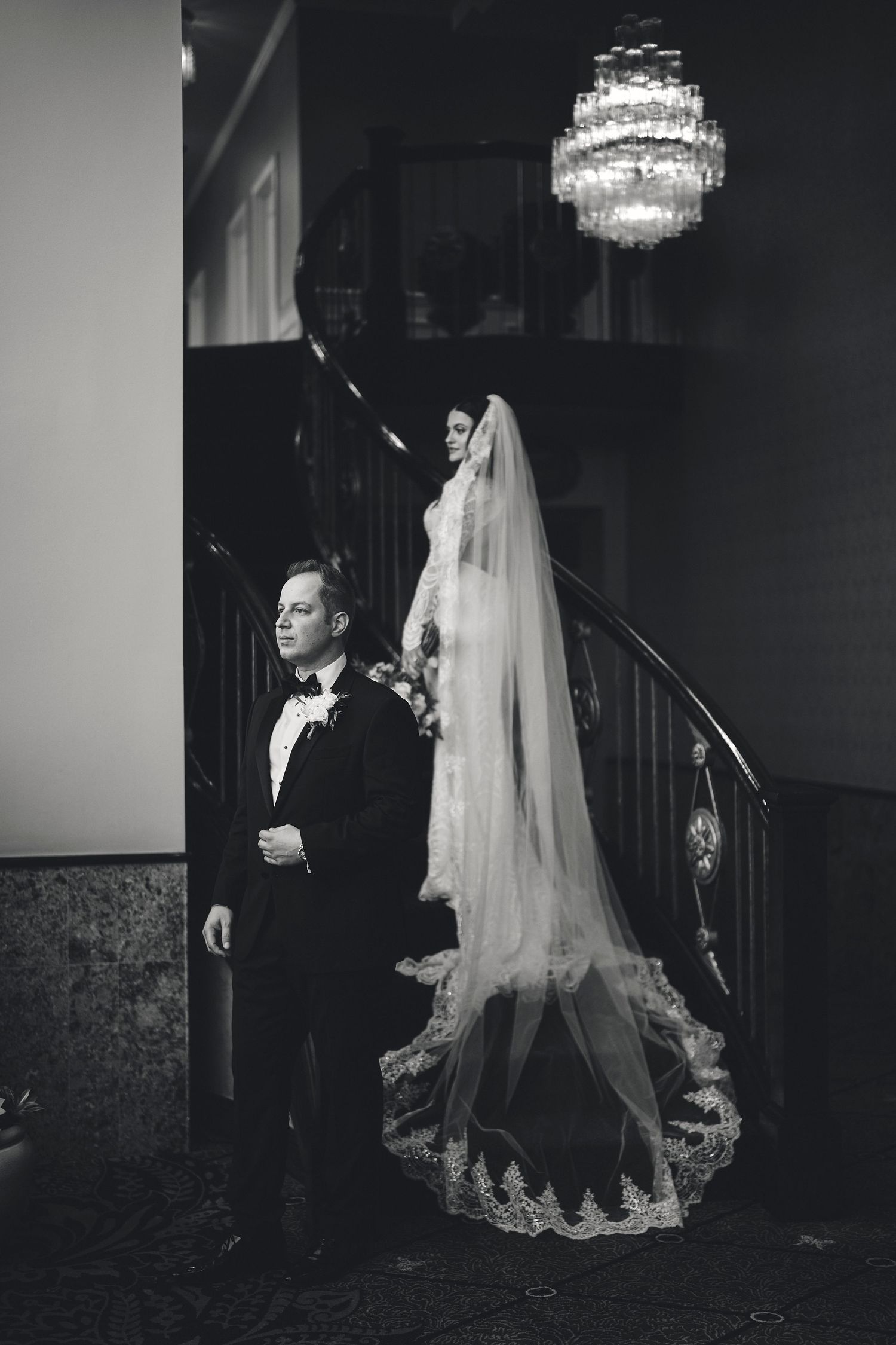 Elegant black and white wedding photo on a grand staircase beneath a crystal chandelier.