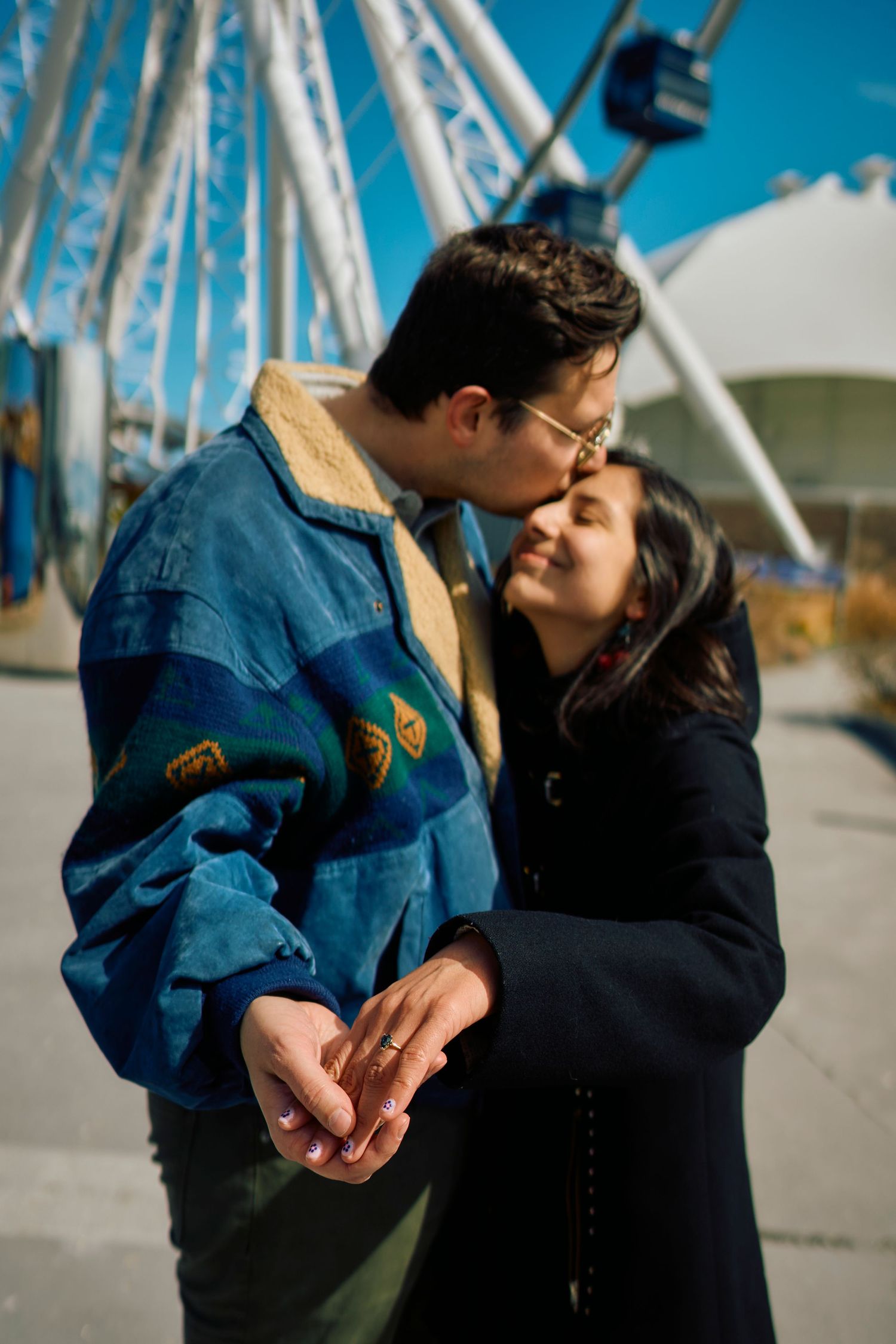 A romantic couple sharing an intimate moment in front of a Ferris wheel on a sunny day at an amusement park.
