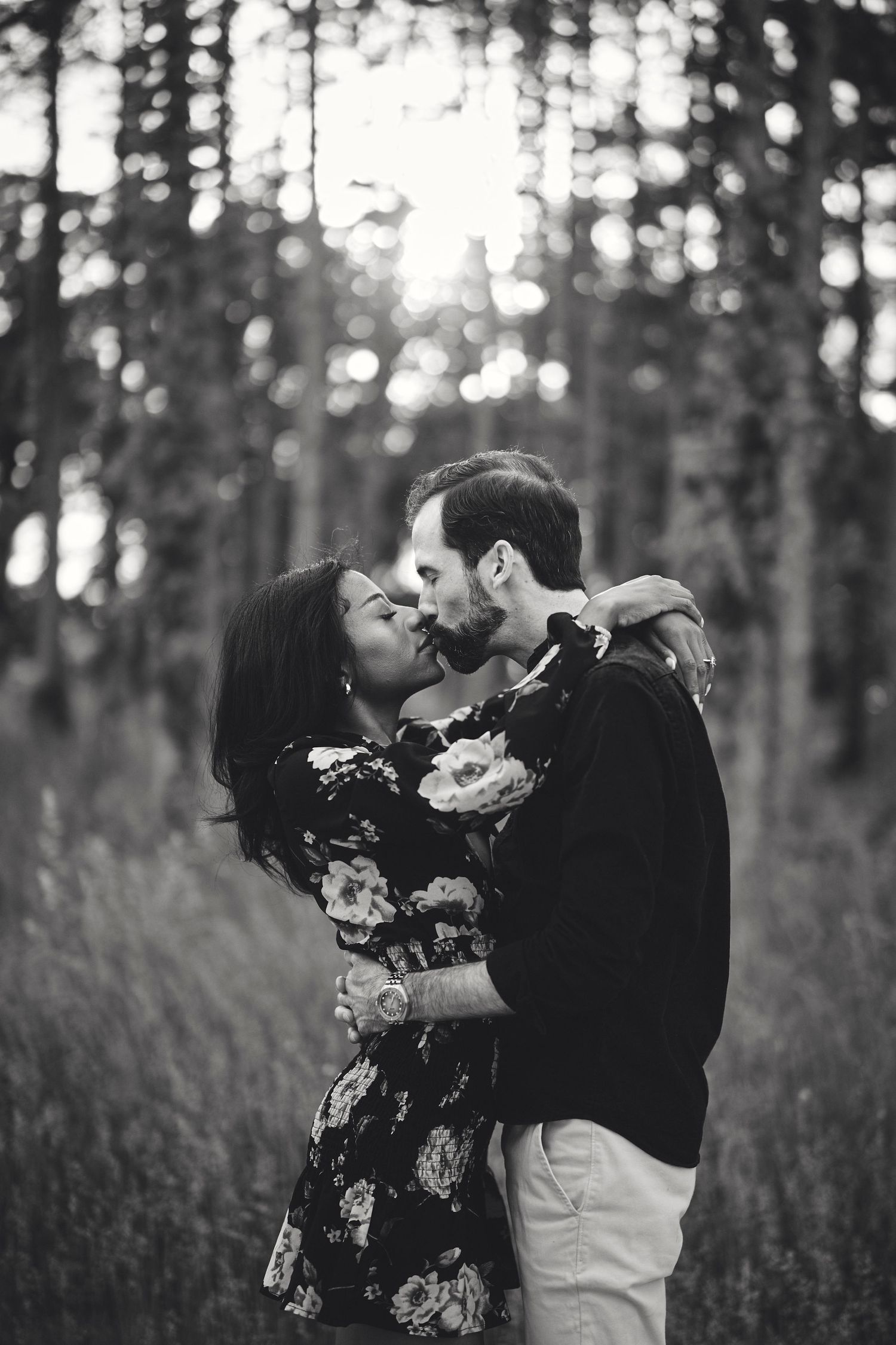 A romantic black and white portrait of a couple embracing in a sunlit forest setting with tall trees in the background.