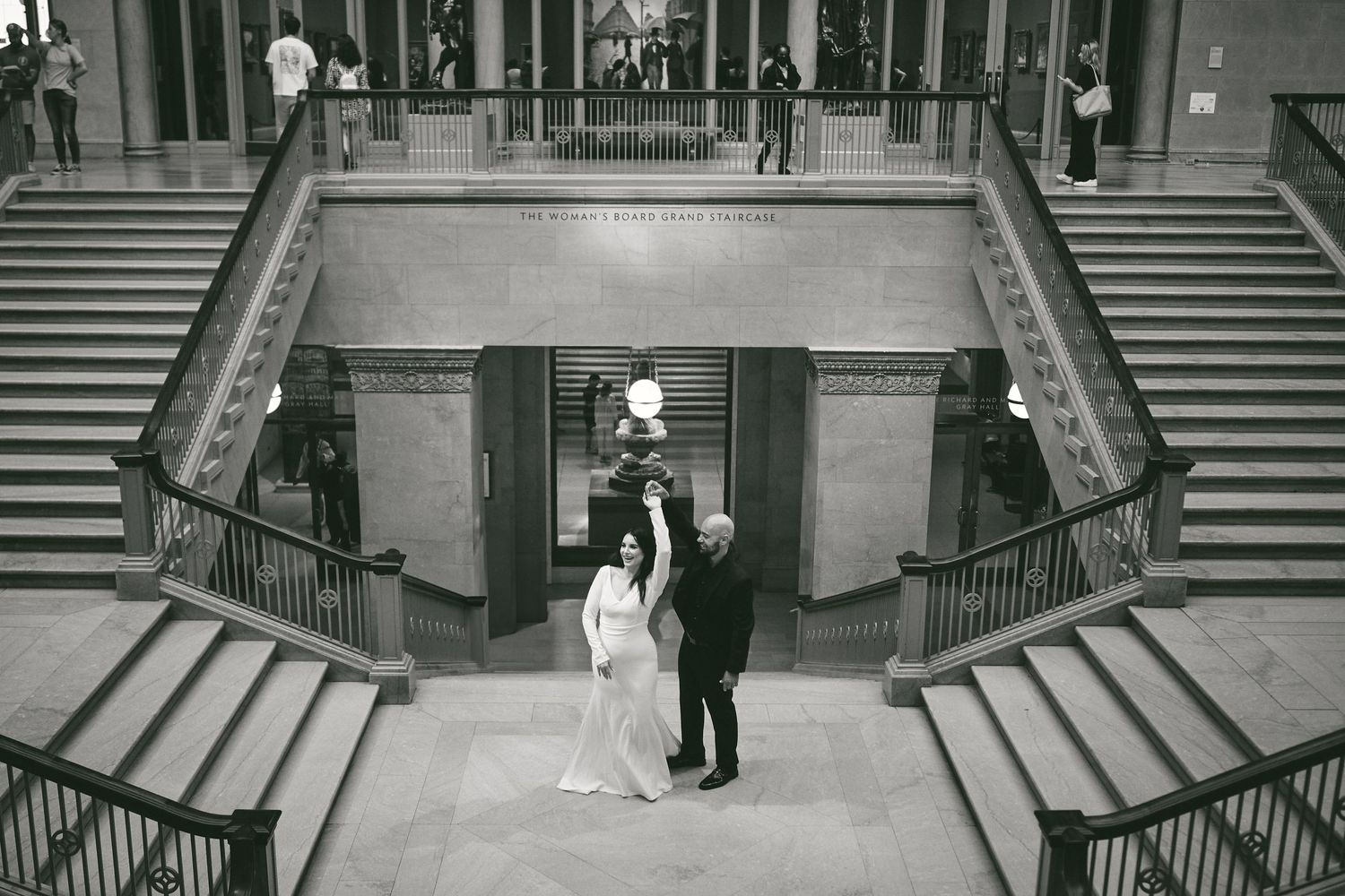 A couple in formal attire stands at the bottom of a grand staircase in black and white photography.