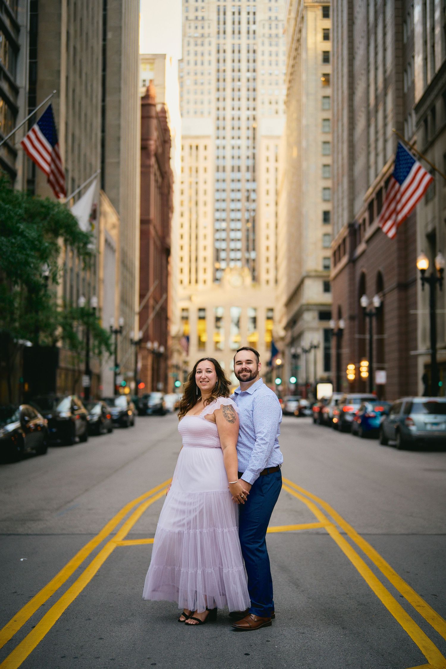 A couple poses elegantly on a downtown city street with tall skyscrapers and American flags in the background.