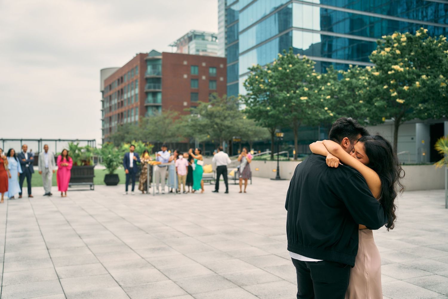 Two figures embrace on an urban plaza while a crowd gathers in the background near modern city buildings.