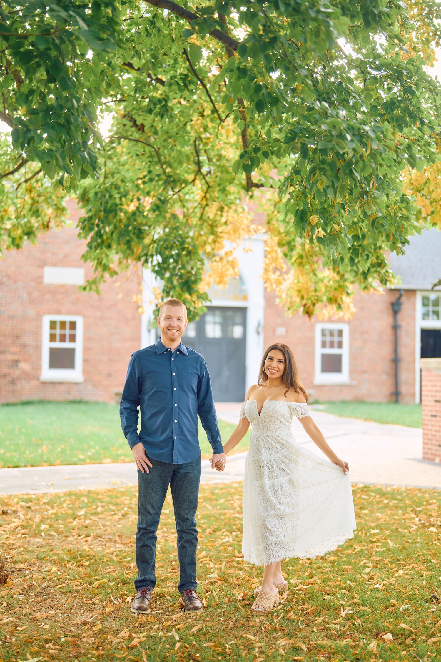 A couple stands together holding hands on fallen autumn leaves in front of a brick building.