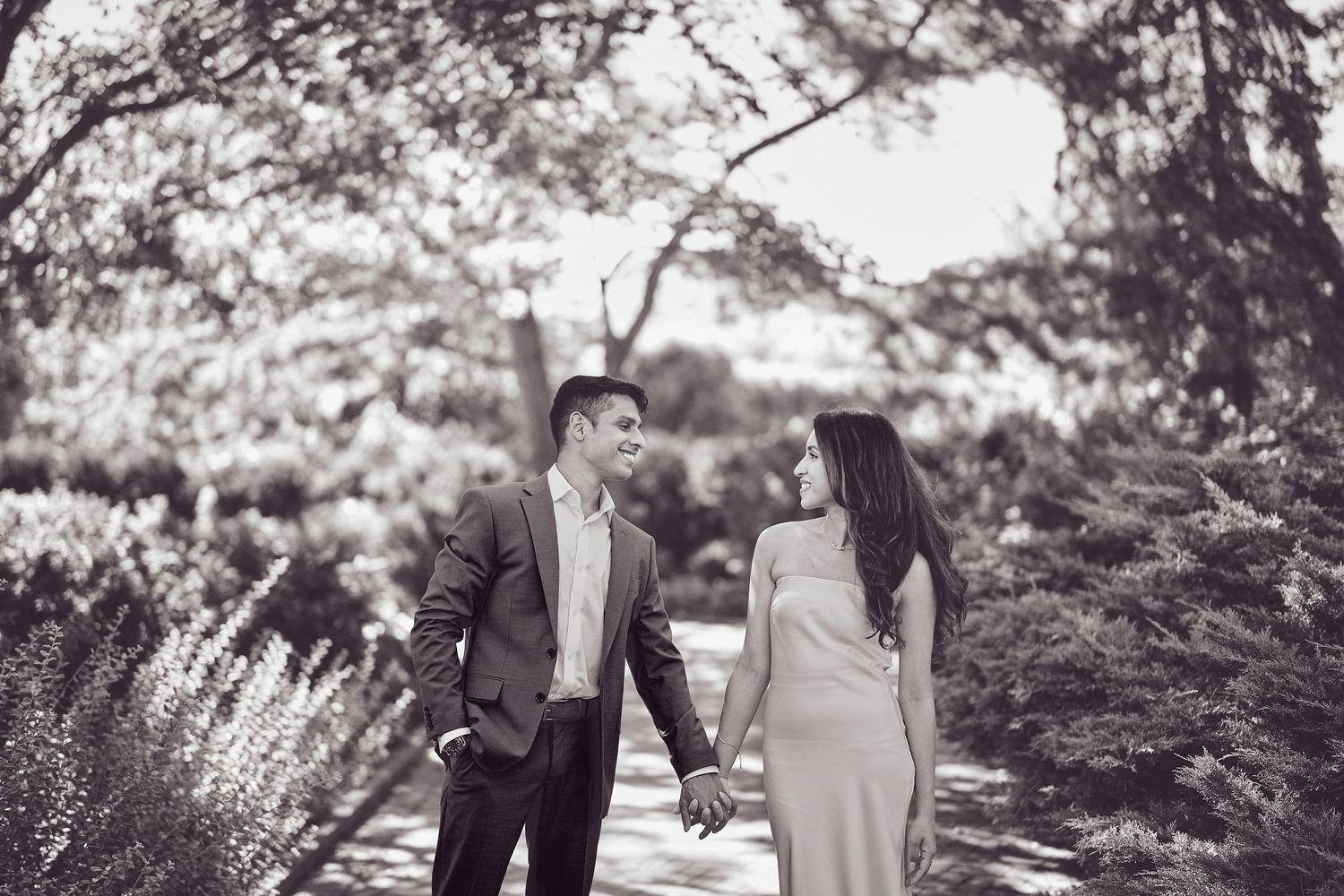 A romantic couple holds hands while walking along a garden path in black and white.