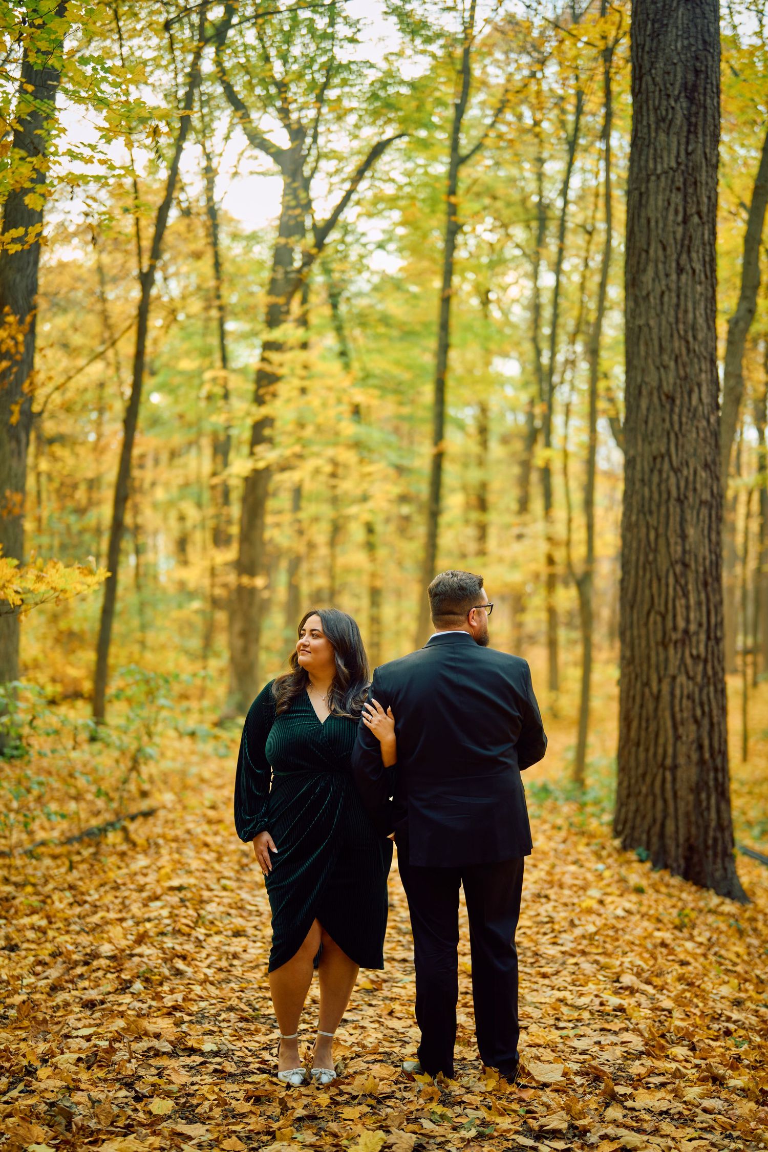A couple walks together on fallen autumn leaves in a forest with golden yellow foliage and tall trees.