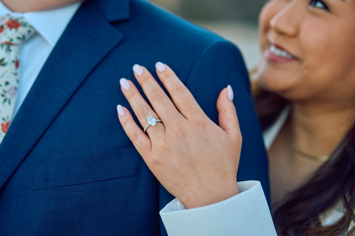 A hand wearing an elegant engagement ring rests on the lapel of a navy blue suit jacket.