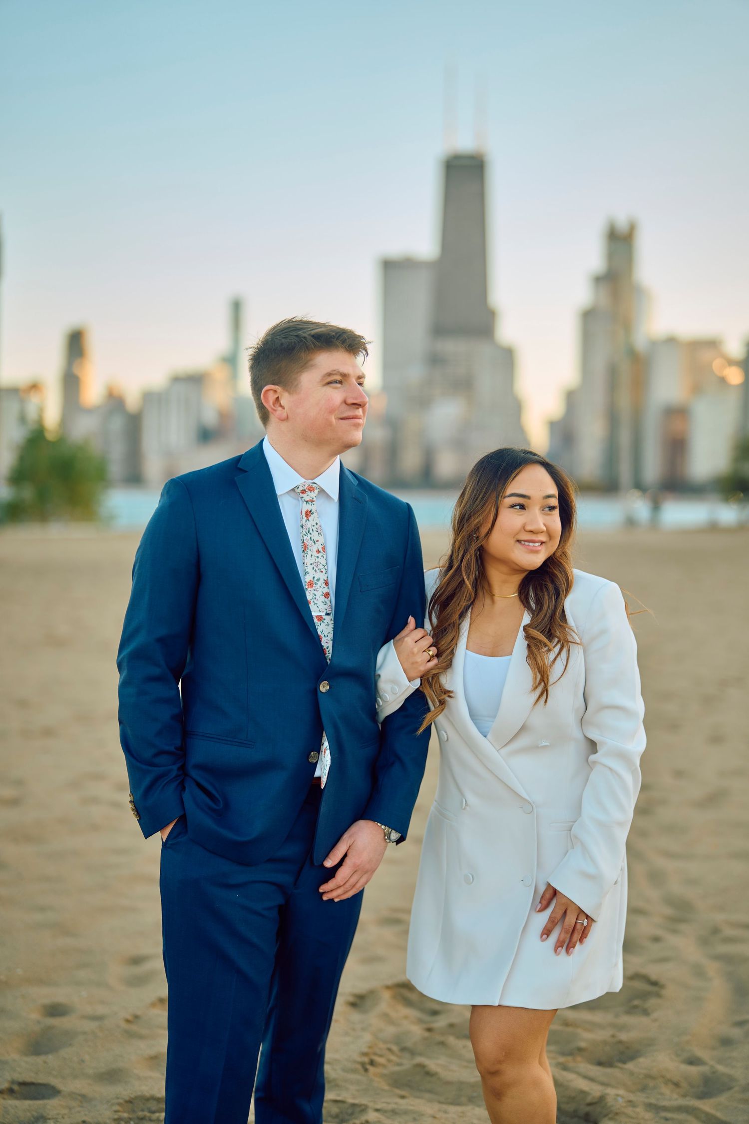 A couple in formal attire poses on a sandy beach with the Chicago skyline visible in the background at sunset.