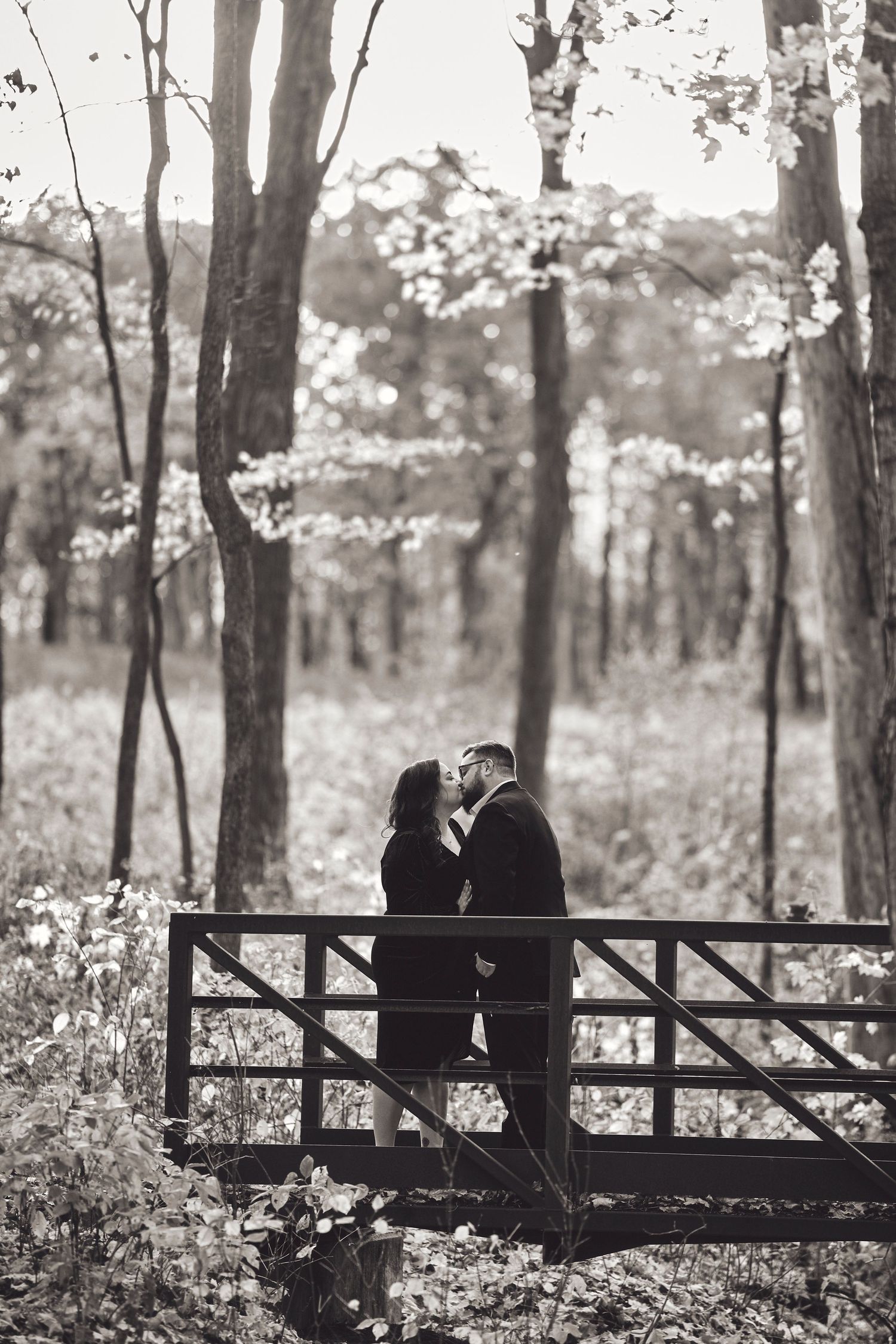 A romantic couple shares an intimate moment while standing on a wooden bridge in a misty forest setting.