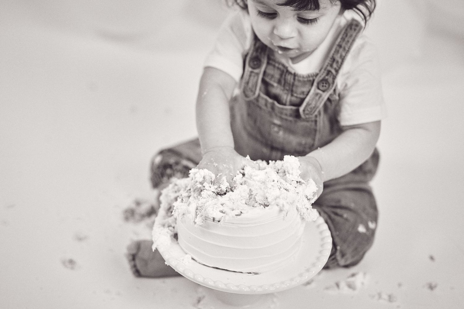 Black and white photo series showing a baby in overalls smashing and eating cake during a first birthday celebration.