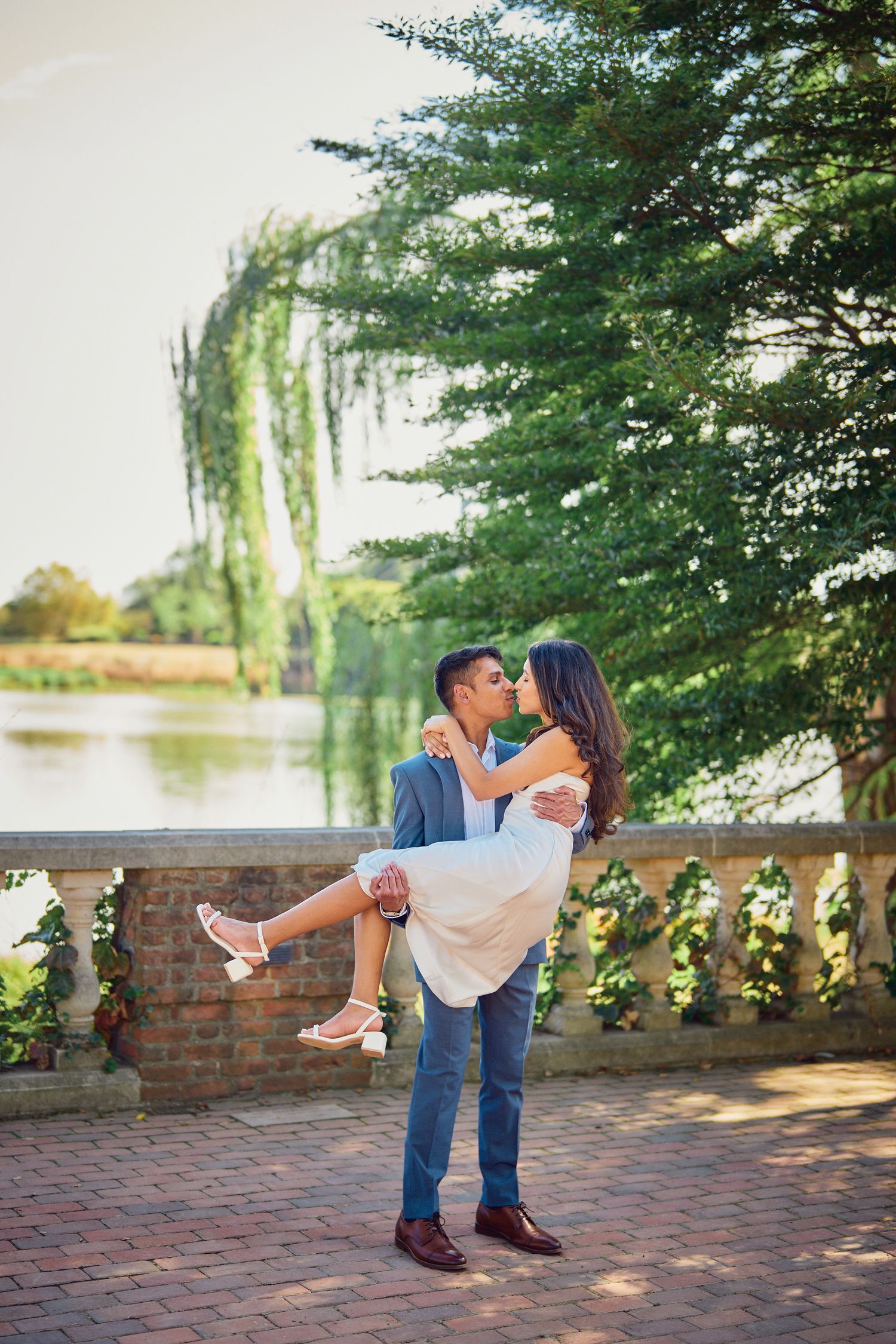 Couple sharing a romantic moment by a lake with weeping willows and brick wall as man carries woman in white dress.