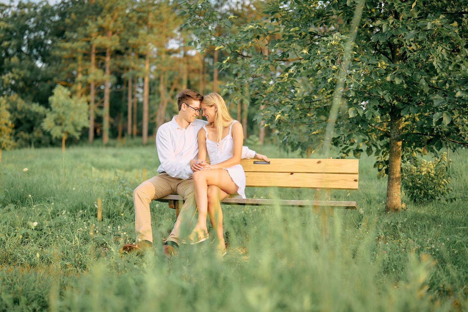 Two people share a romantic moment on a wooden bench surrounded by lush green trees and grass in a sunlit garden setting.