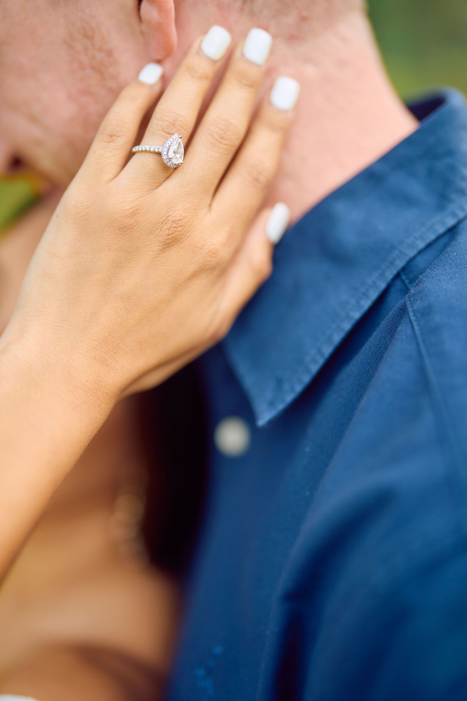 Close-up of a hand with white manicured nails showing off an oval diamond engagement ring against a navy blue shirt.