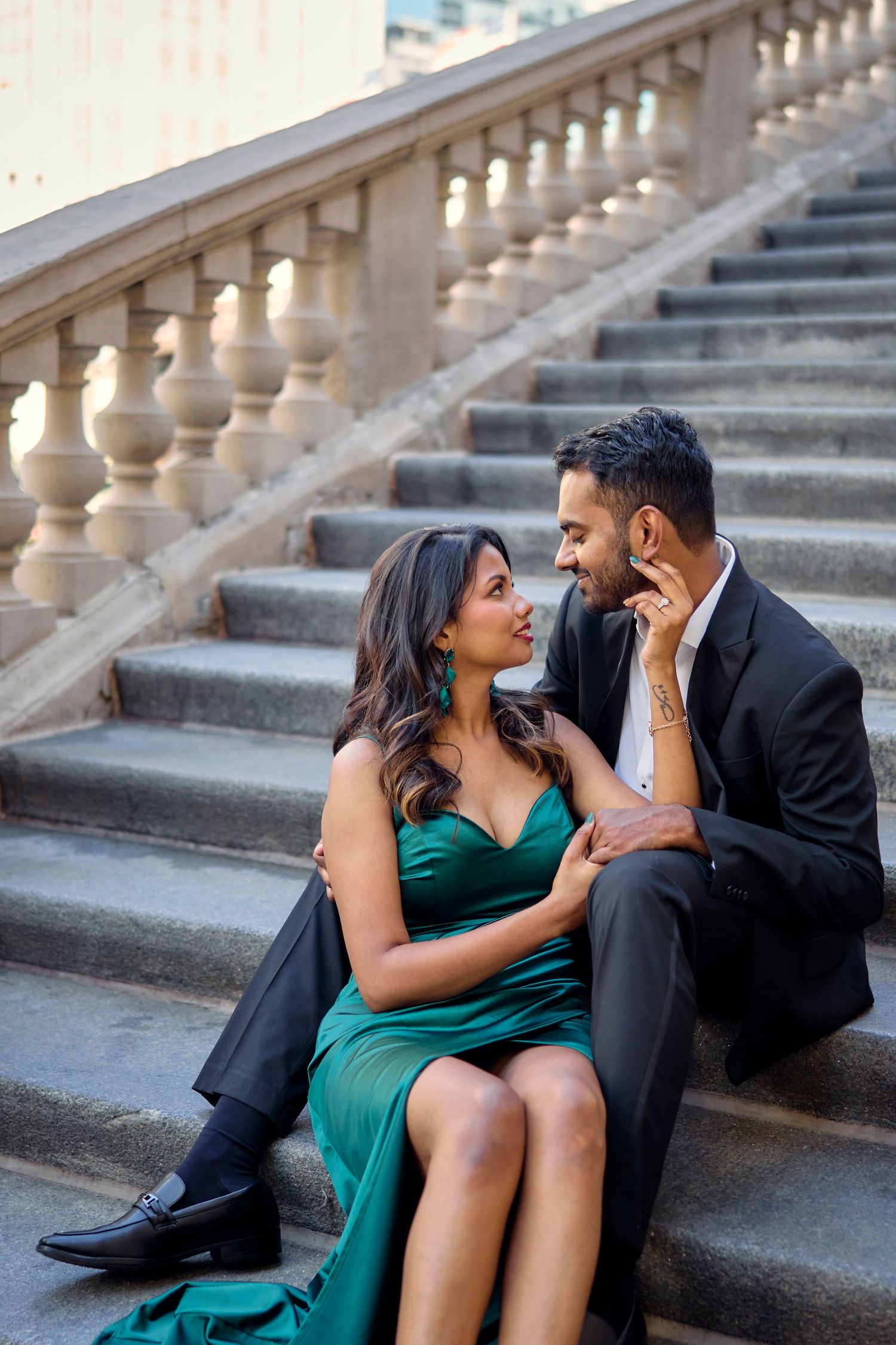 A couple share an intimate moment on elegant stone stairs, with the woman in emerald green and the man in black suit.