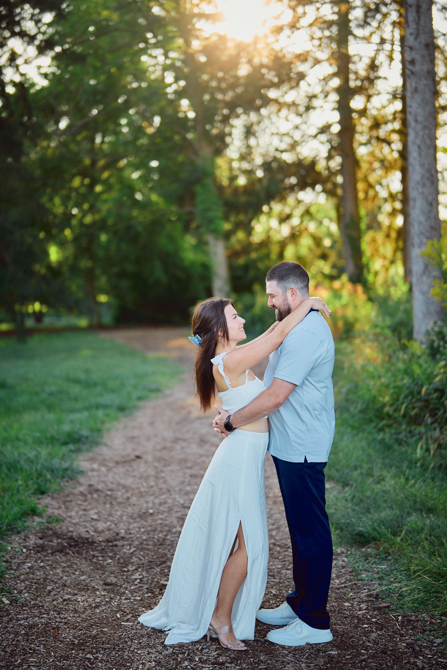 A romantic couple embraces on a sunlit forest path during golden hour with trees and greenery in the background.