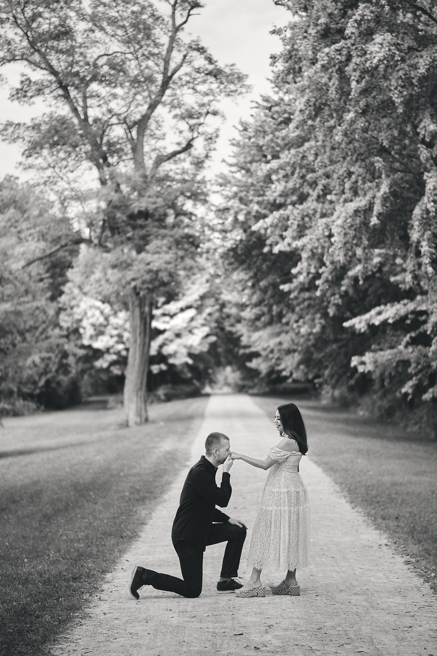 A romantic proposal moment captured in black and white on a tree-lined path.