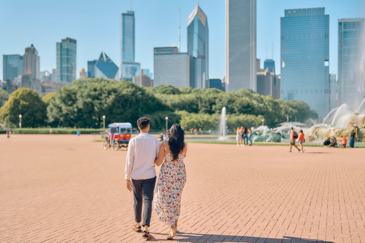A couple walks together on the red brick pavers at Millennium Park with Chicago's skyline towering in the background.