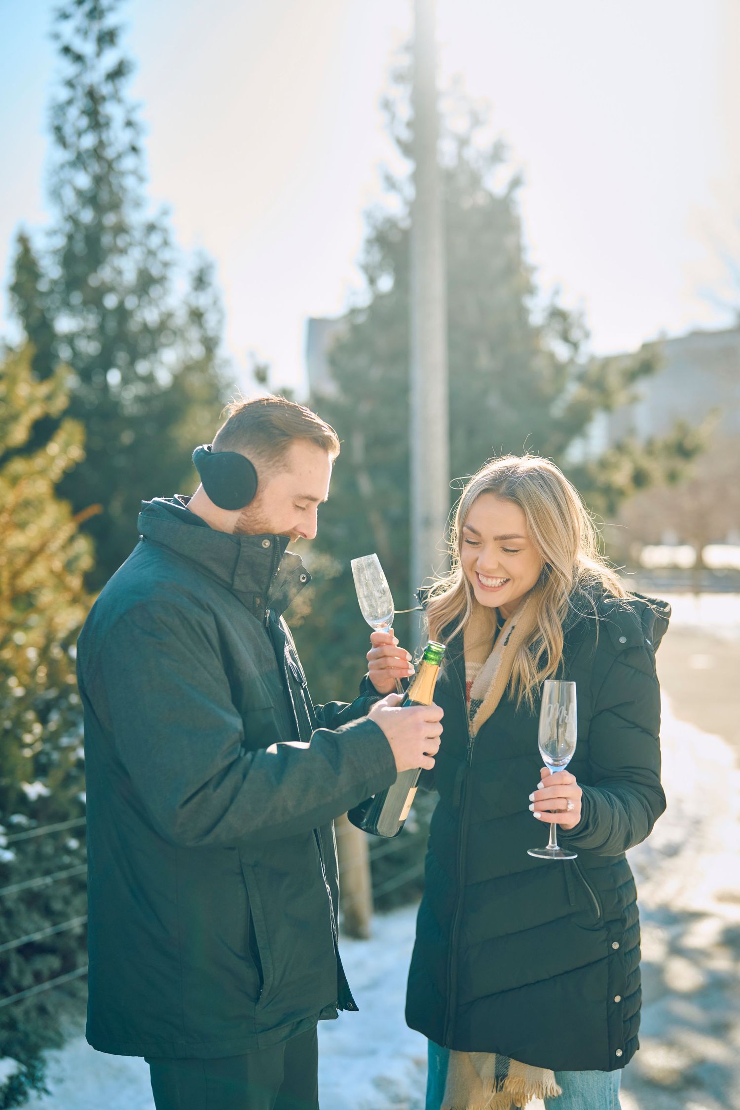 Two people in winter coats enjoying champagne outdoors in a snowy setting with evergreen trees in the background.