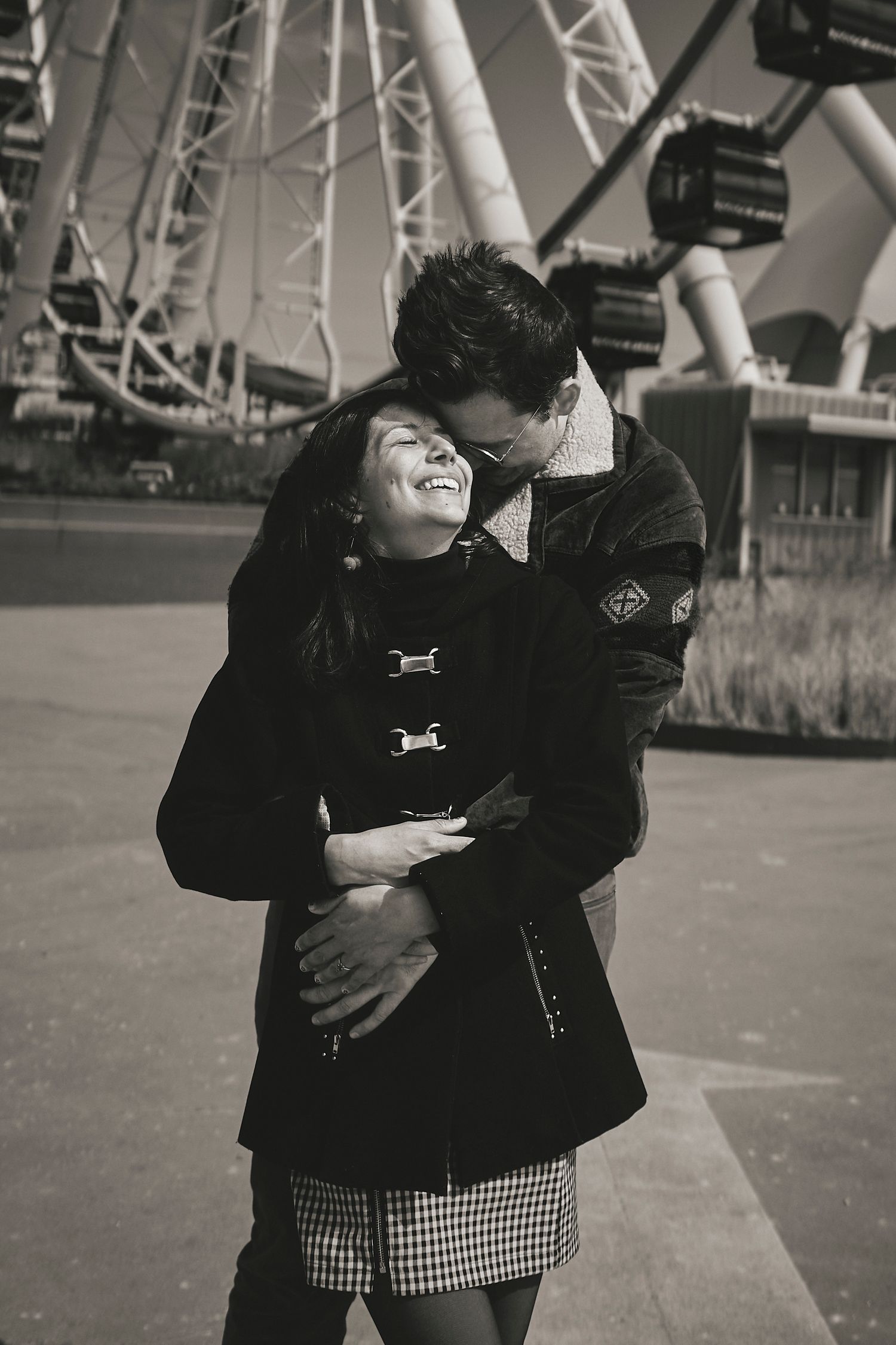 Black and white romantic moment captured in front of a ferris wheel as a couple embraces and shares a joyful laugh together.