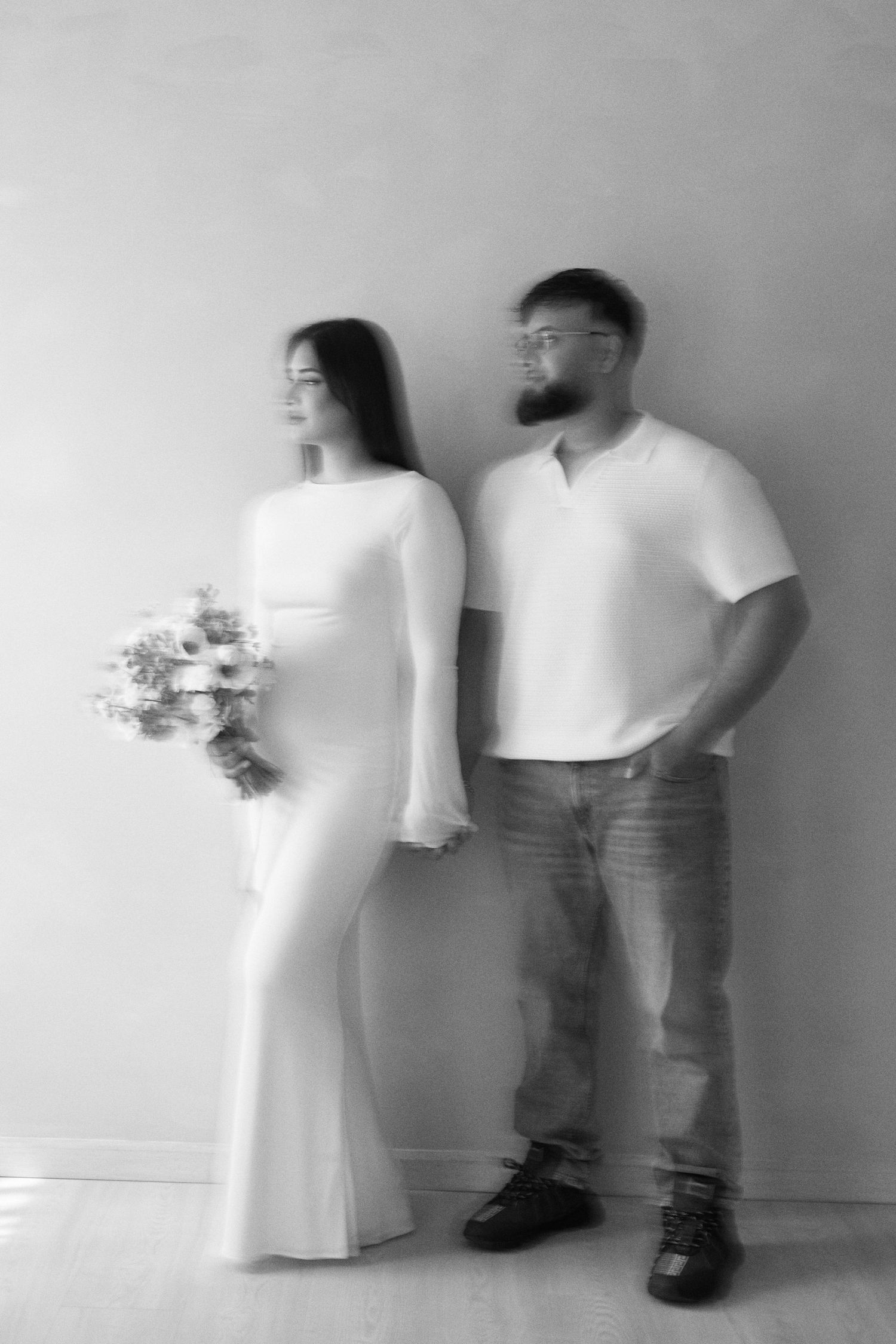 A black and white artistic portrait of a couple in white outfits standing against a plain wall, with a floral bouquet.