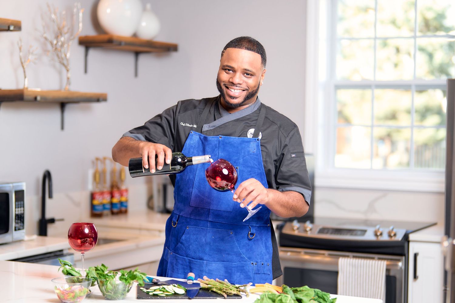 A person in a blue apron prepares ingredients in a bright modern kitchen with wooden shelves.