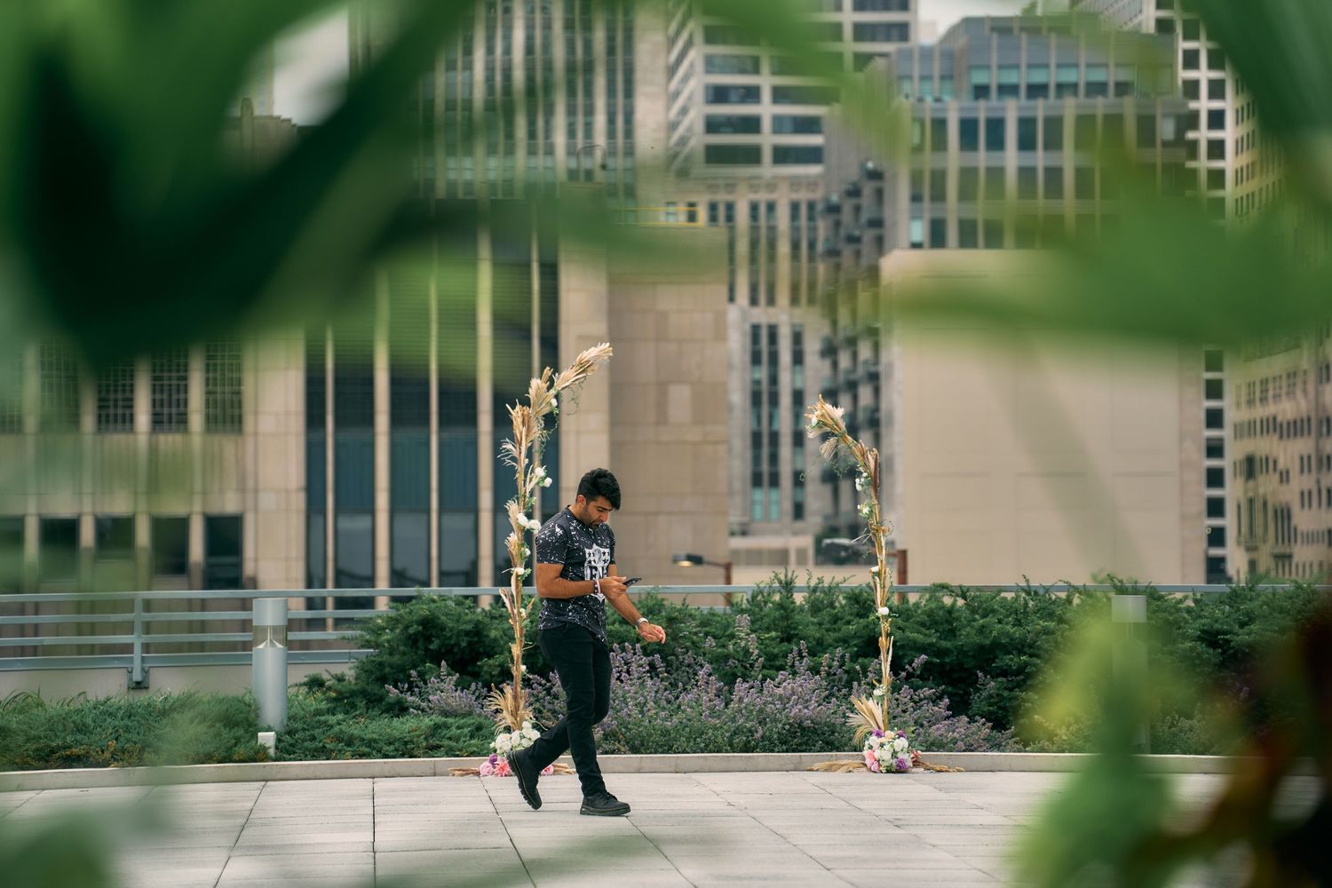 A person in a dark uniform walks past decorative light posts along a city path with blurred greenery in the foreground.